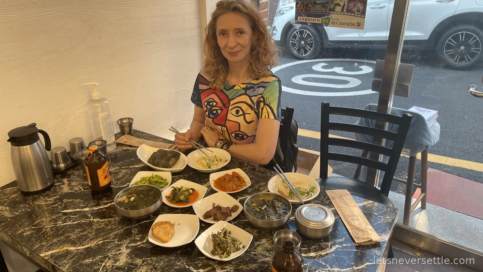 Inga at a small marble table crowded with banchan side dishes in a Busan eatery, the street visible outside