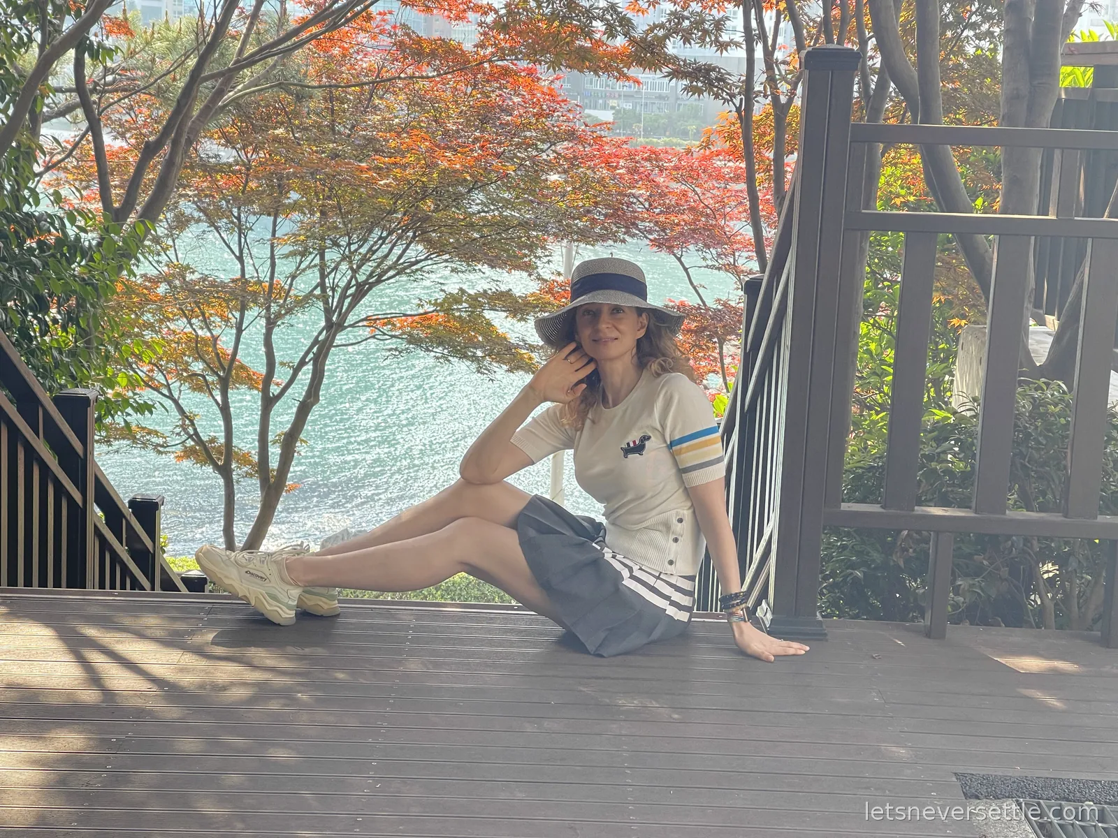 Inga in a sun hat sitting on wooden boardwalk steps above turquoise sea, framed by red maple leaves