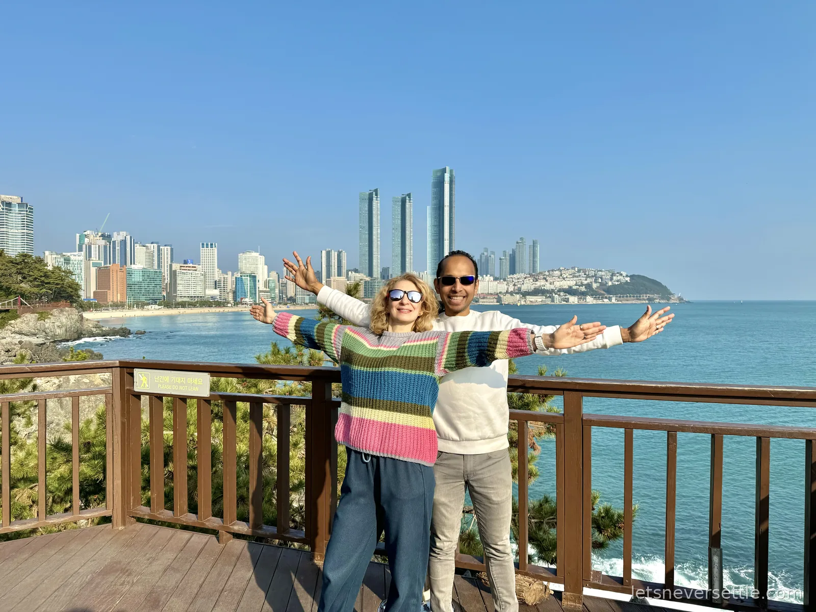 Inga and Dileep with arms spread wide on a wooden deck on Dongbaekseom, Haeundae's skyscrapers across the bay