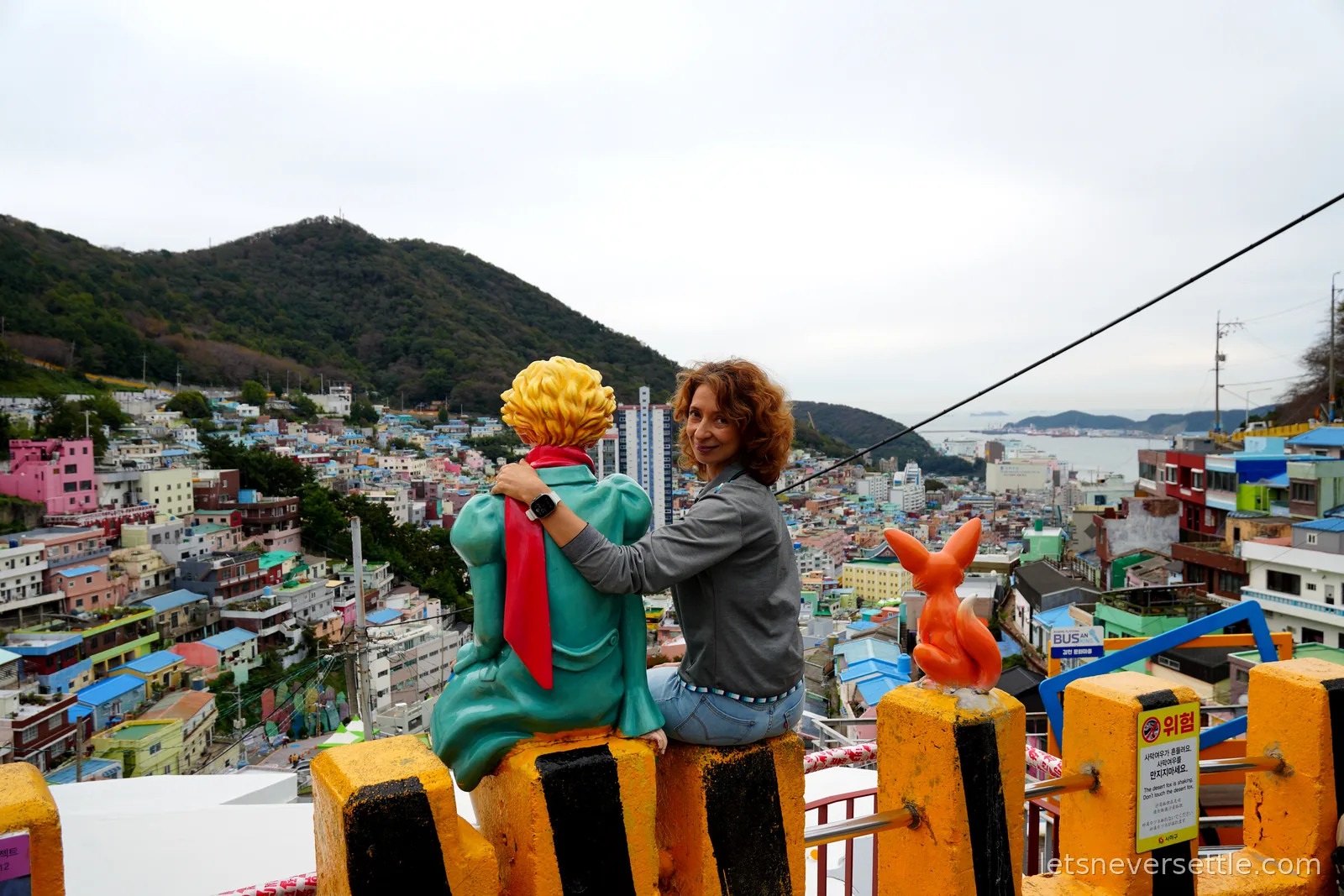 Inga sitting beside the Little Prince statue on a yellow railing above Gamcheon Culture Village, the fox statue and the port in the distance