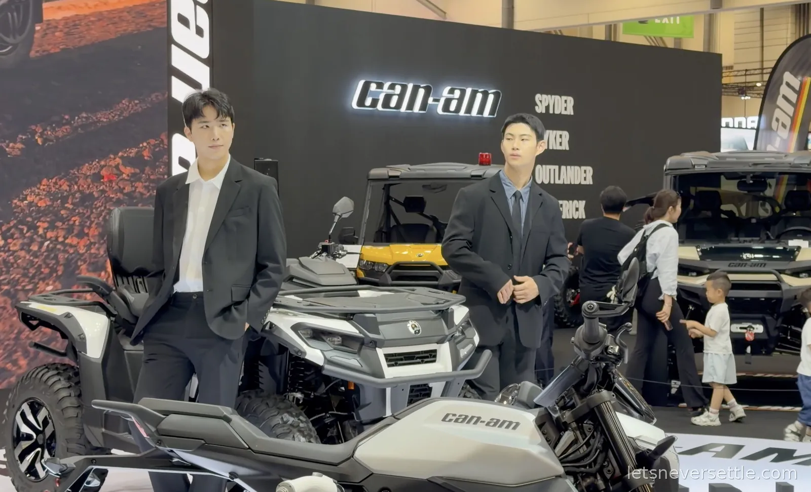 Two young male booth models in black suits standing between Can-Am Outlander ATVs at the Busan Mobility Show, the stand wall behind them lettered Spyder, Ryker, Outlander and Maverick