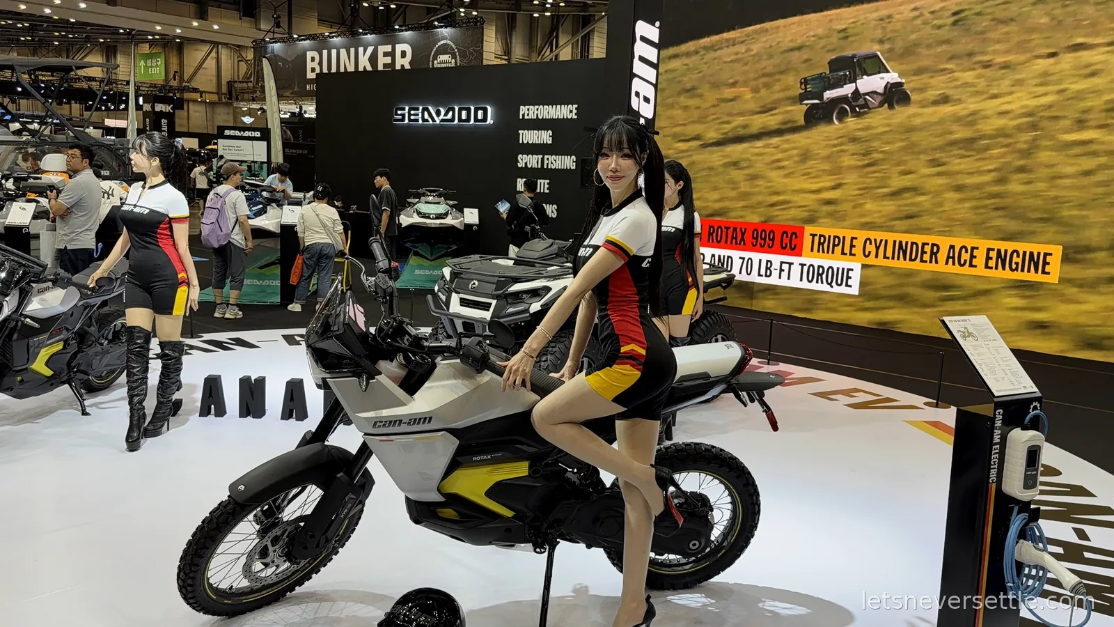 A white Can-Am electric motorcycle on a display stand at the Busan Mobility Show with a Sea-Doo stand and an off-road buggy on the screen behind