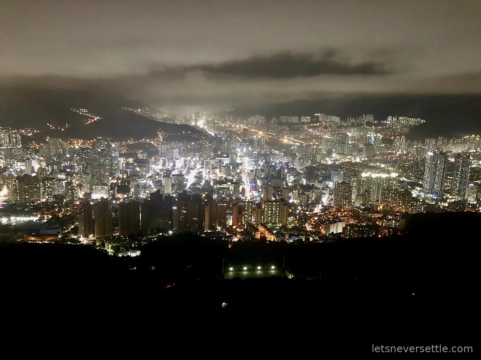 Busan city lights at night seen from a mountain viewpoint, with low cloud hanging over the hills