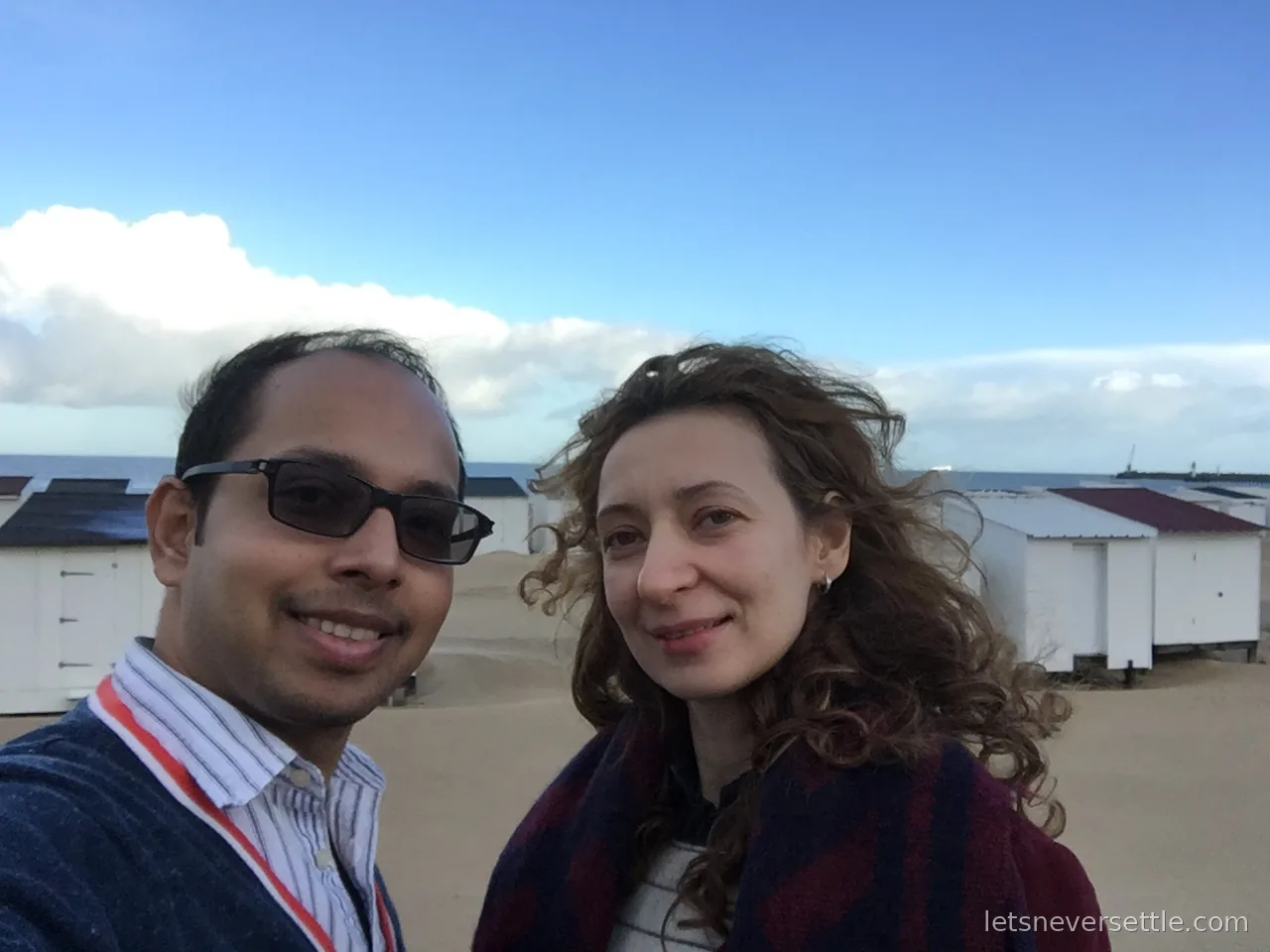Inga and Dileep taking a selfie on Calais beach in front of white beach huts, winter dunes and the sea