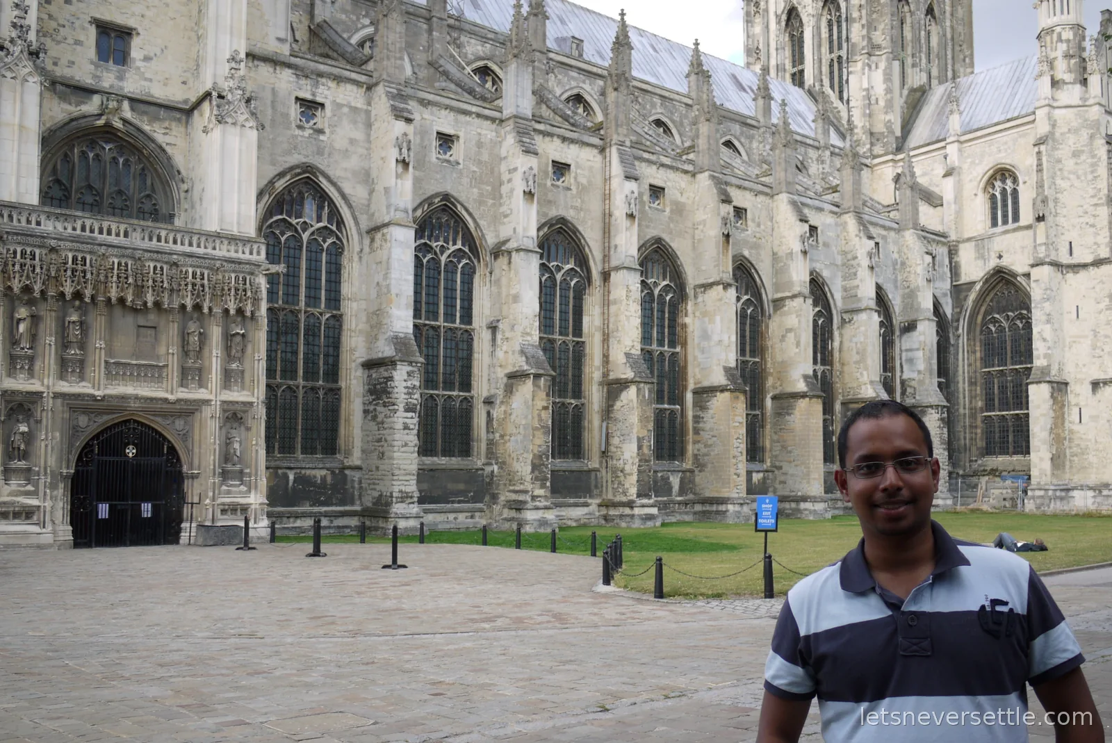 Dileep standing in the precincts beside the long stone flank of Canterbury Cathedral, with a blue sign pointing to the shop, exit and toilets