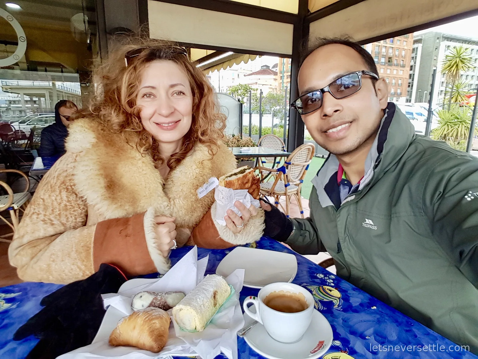 Inga and Dileep at a cafe table with a blue patterned cloth, eating pastries and a filled roll with an espresso in front of them and palm trees outside