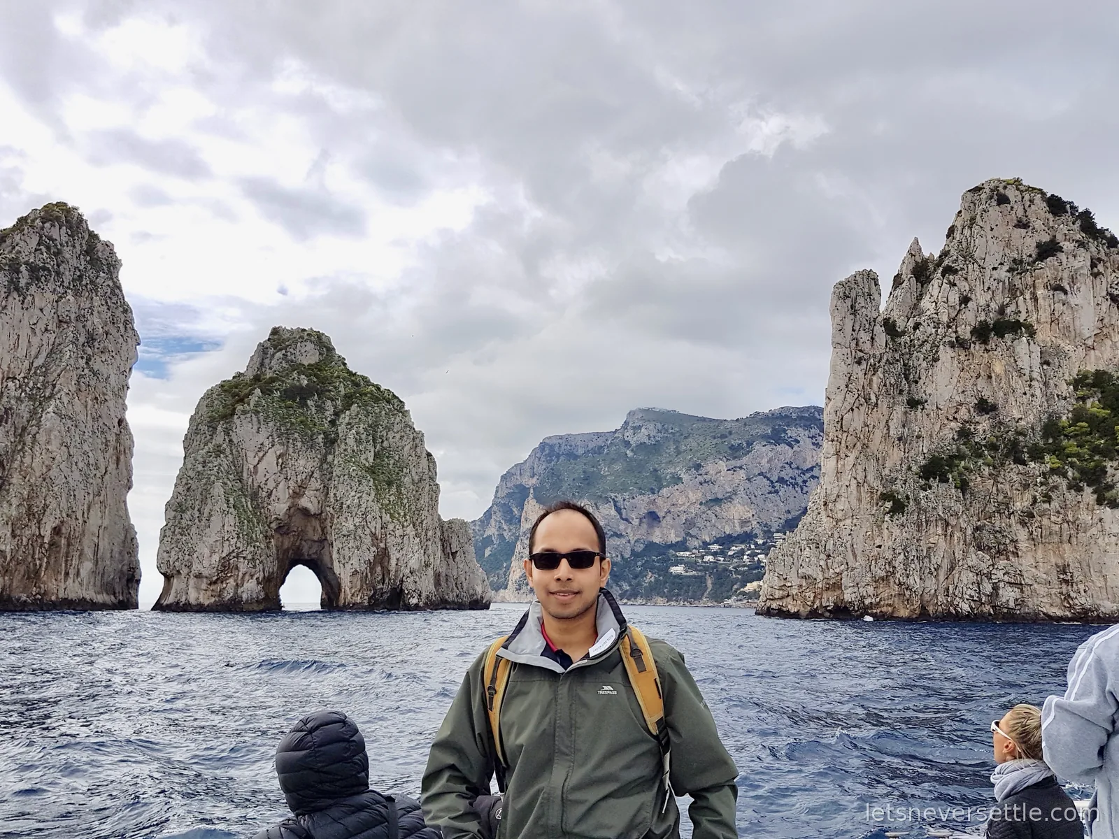 Dileep on the deck of a boat with the three Faraglioni rock stacks rising out of the sea behind him, the middle one pierced by a natural arch