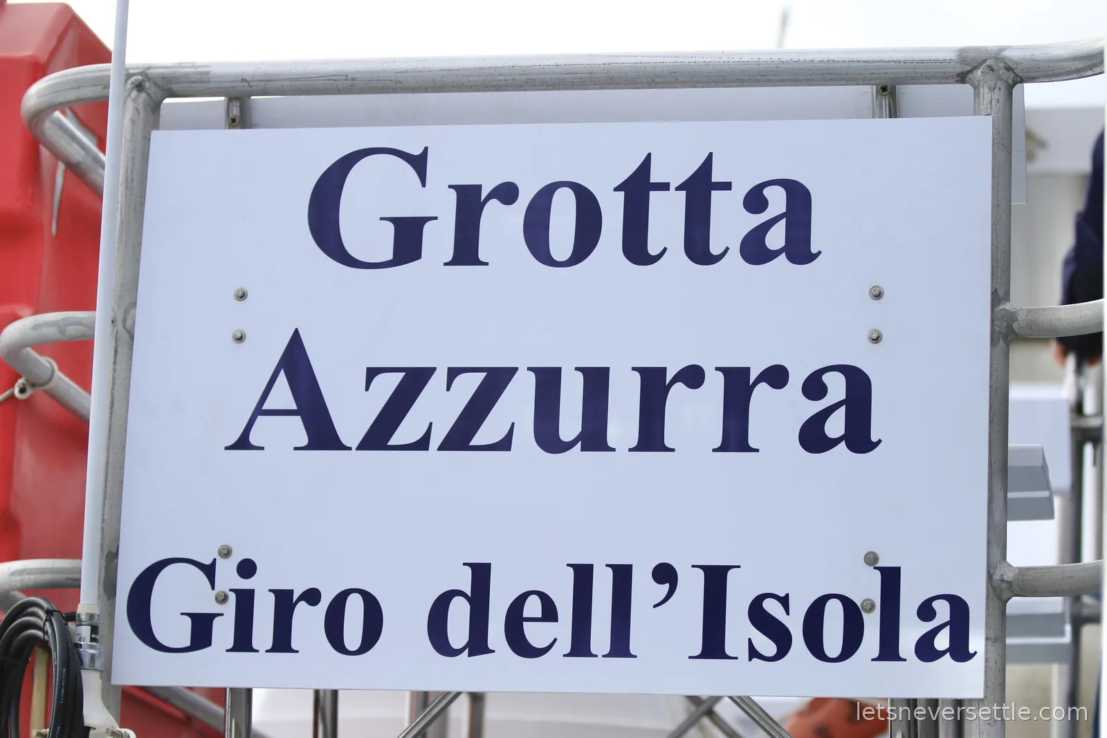 A white sign lettered Grotta Azzurra, Giro dell'Isola, bolted to the metal railing of a boat