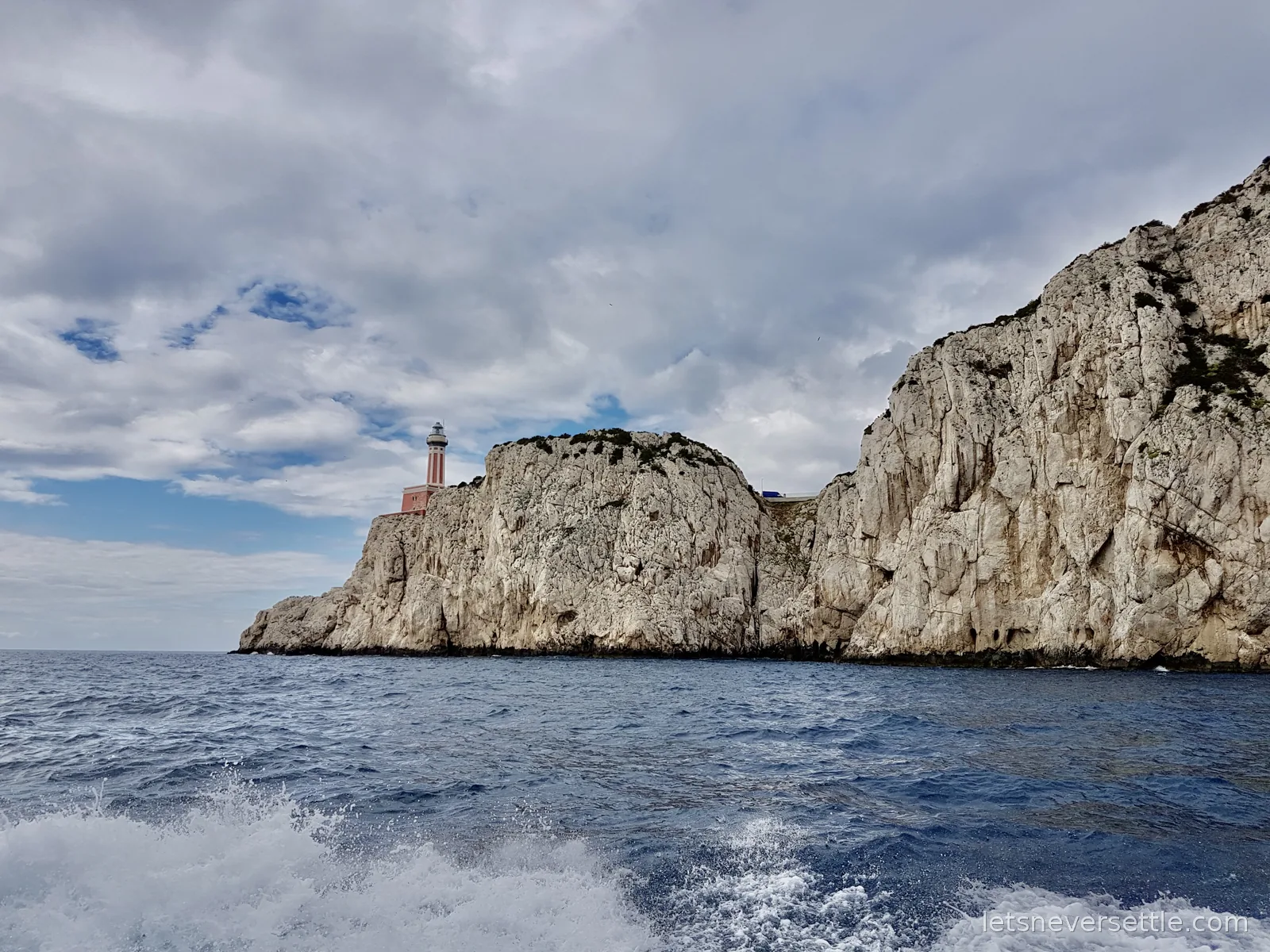 A red and white striped lighthouse standing on top of pale limestone cliffs above a choppy blue sea, seen from a moving boat
