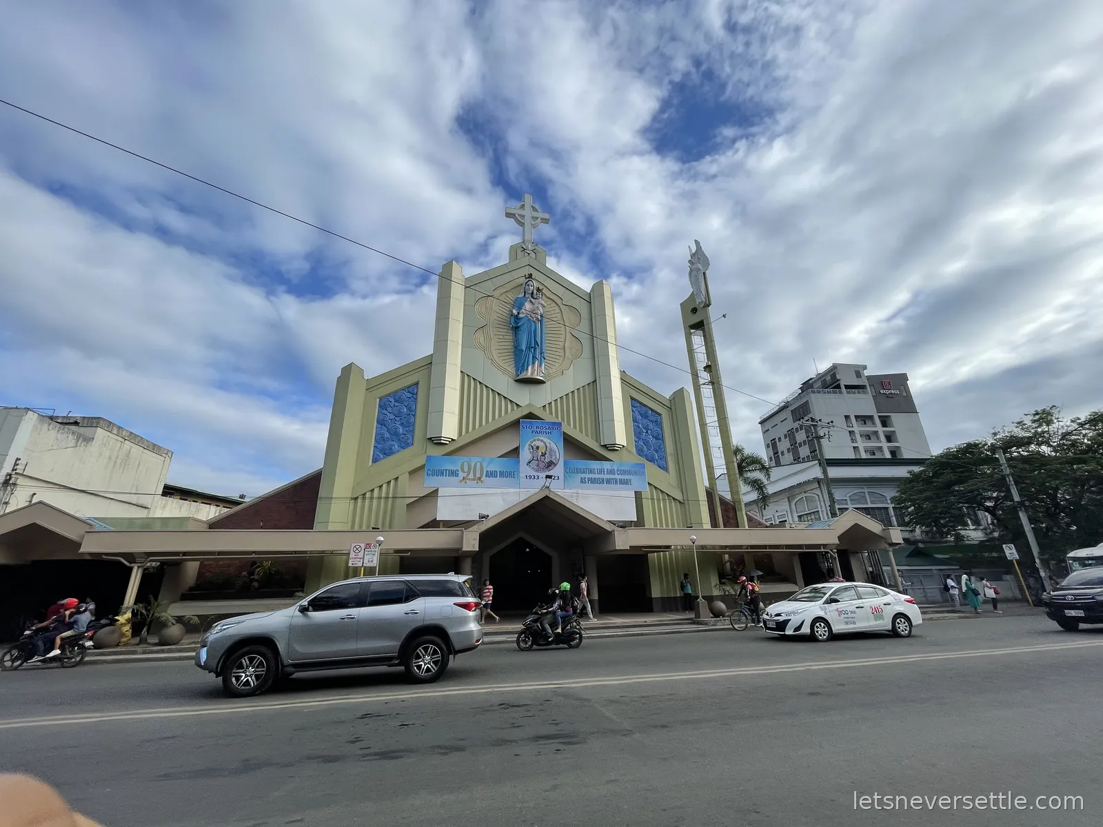 A parish church in Cebu City with a pale green and cream facade, a statue in a niche above the doors and a cross on the bell tower, cars parked outside