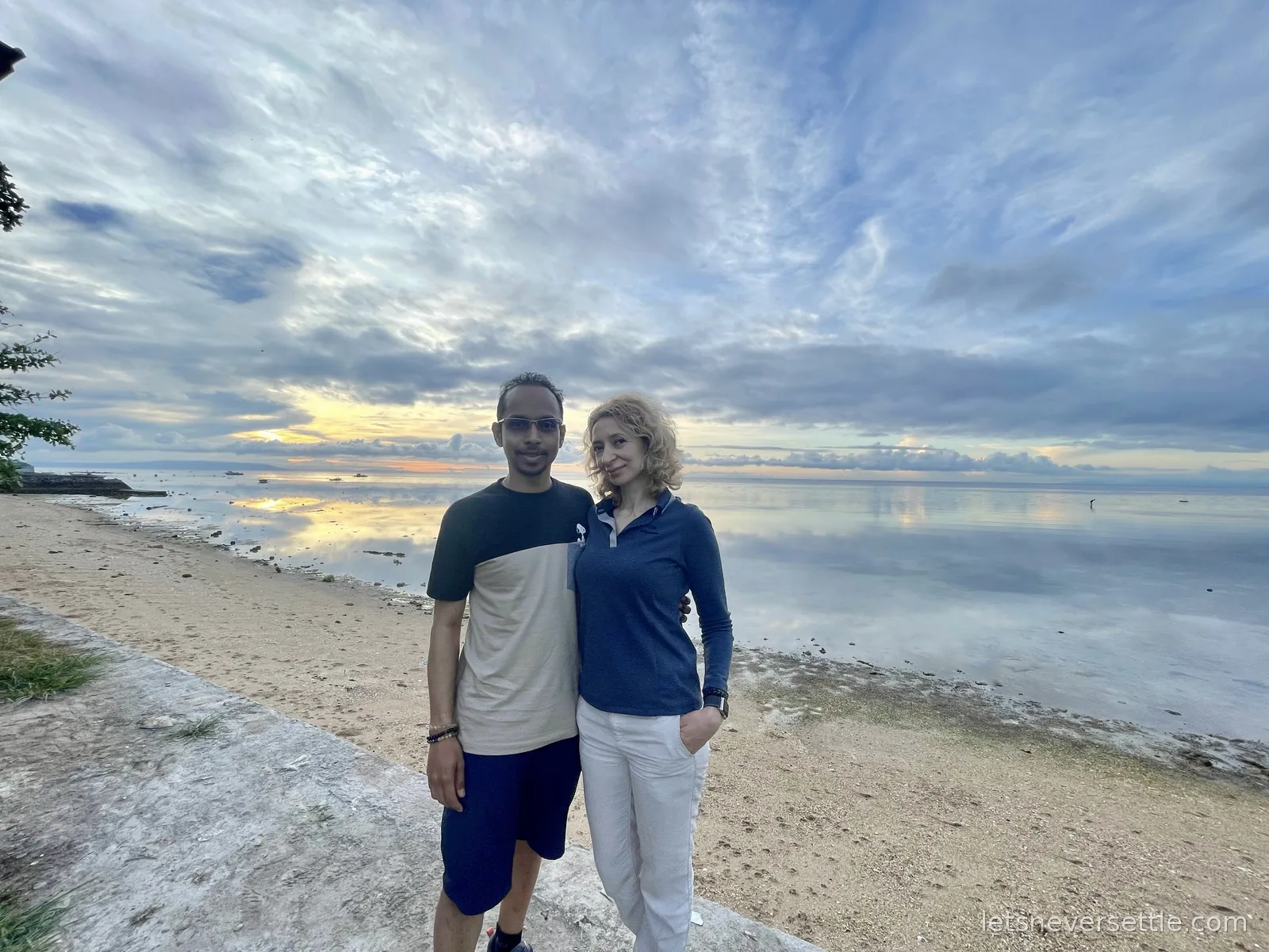 Inga and Dileep standing on a sea wall at sunrise with a mirror-calm sea and small fishing boats behind them