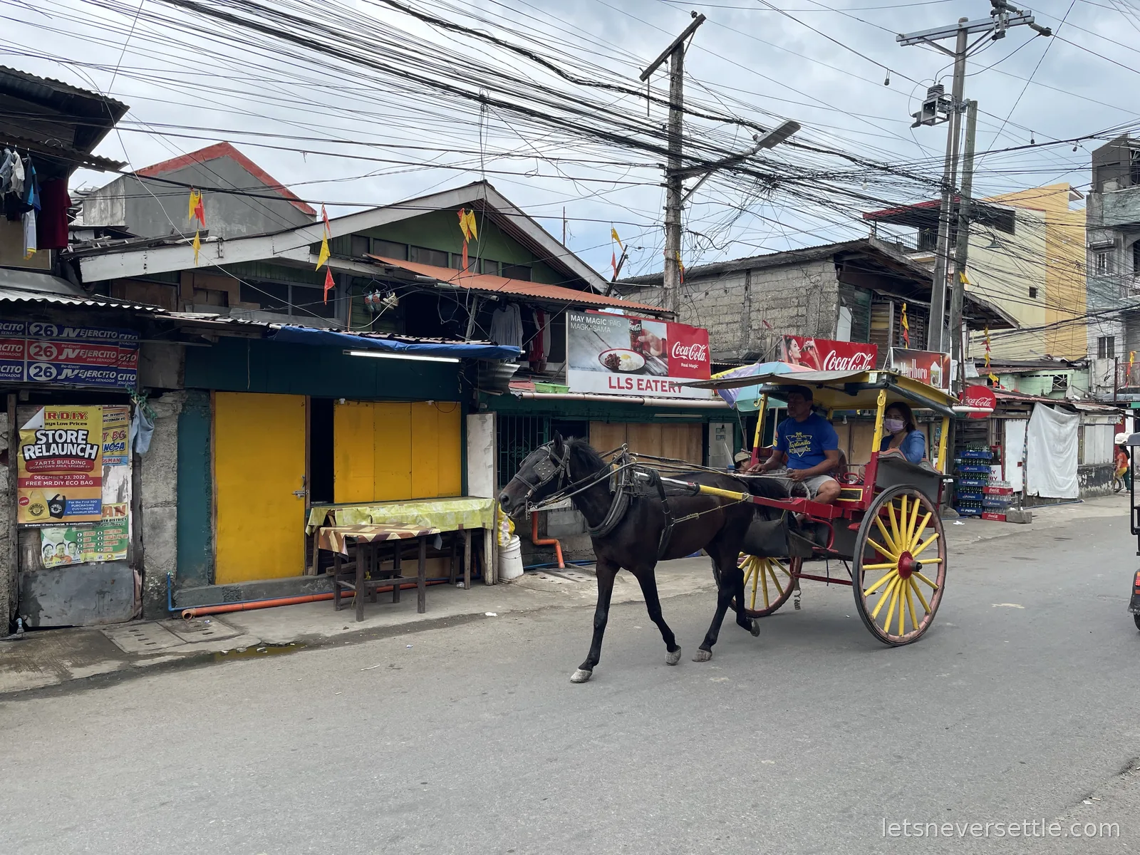 A horse-drawn tartanilla with yellow-spoked wheels passing shuttered shopfronts under a dense tangle of overhead cables in Cebu City