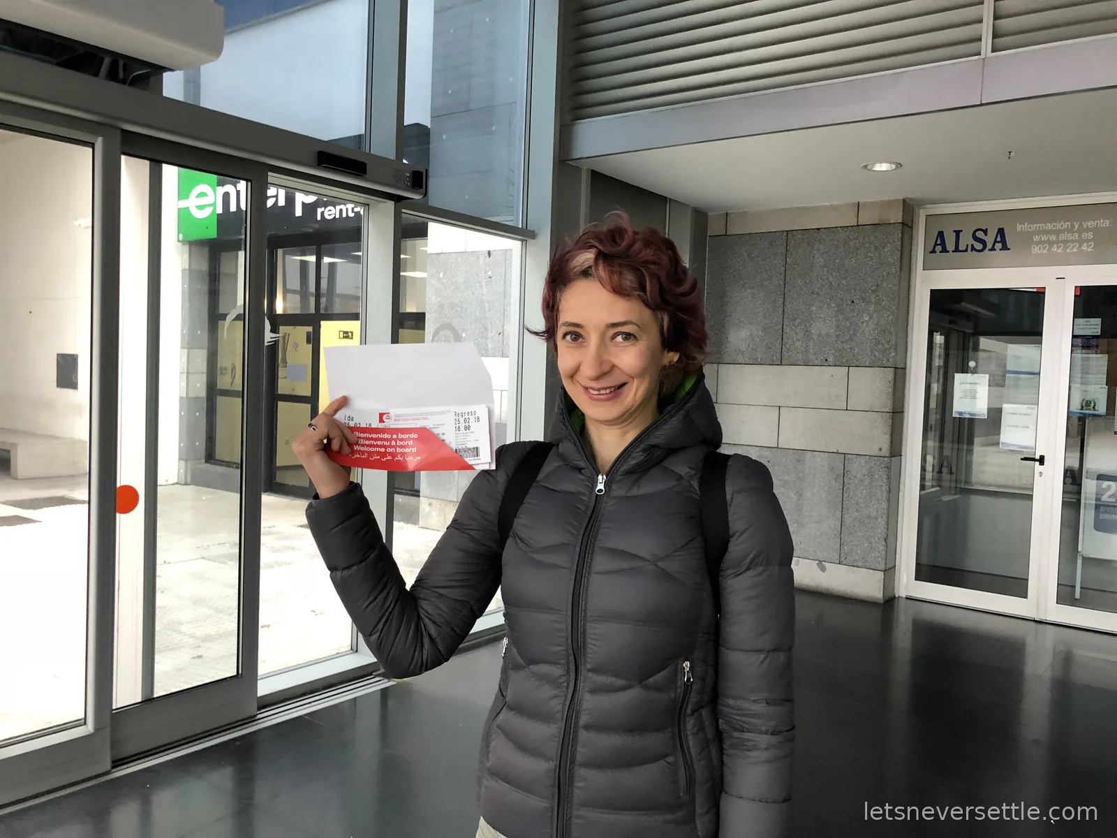 Inga at the Algeciras terminal holding a red ferry ticket wallet printed Welcome on board in four languages, an ALSA office behind