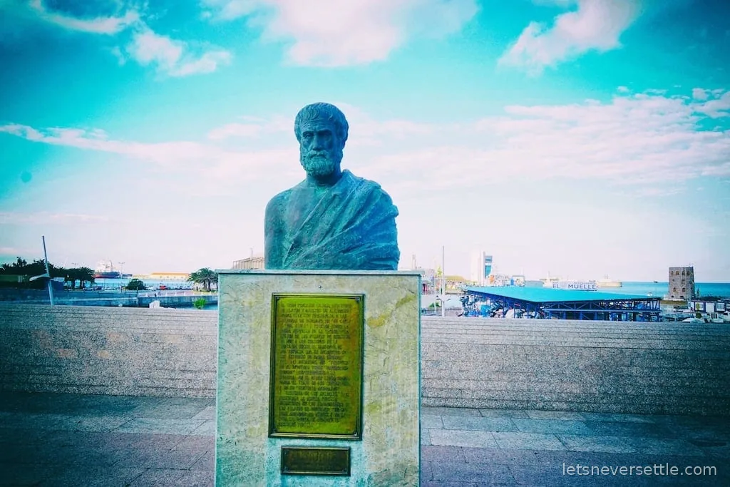 A bronze bust of Aristotle on a marble plinth by the seafront, with the marina behind