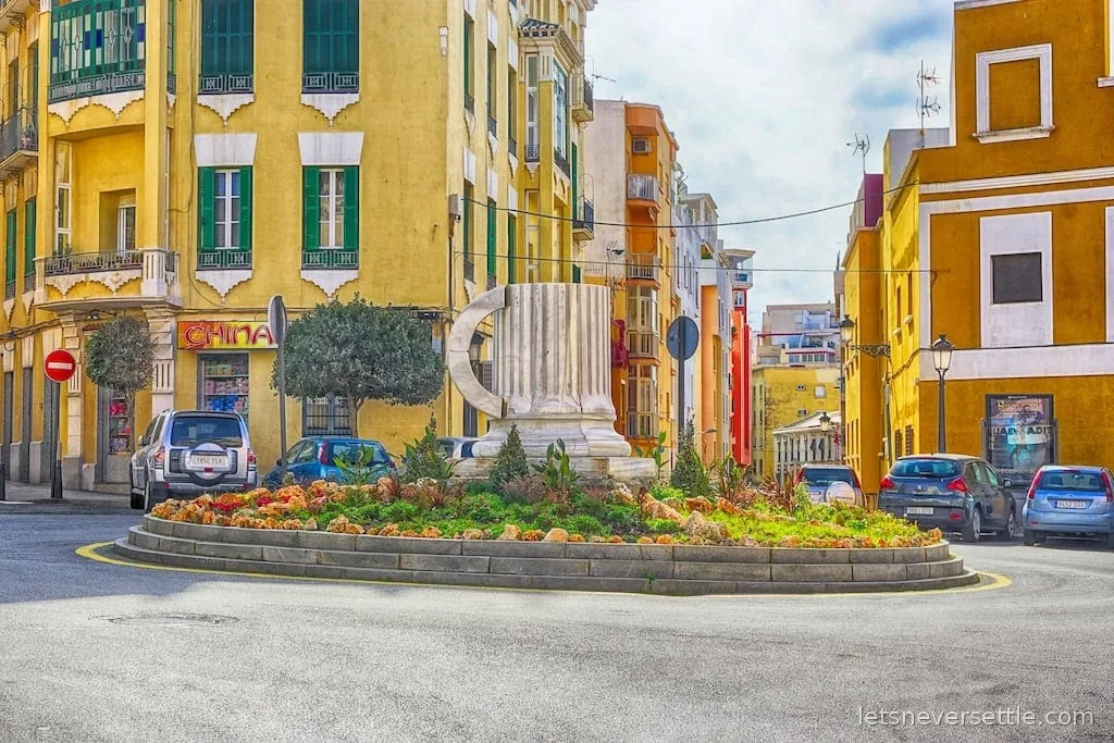 A broken marble column monument on a flowered roundabout surrounded by ochre-yellow colonial buildings