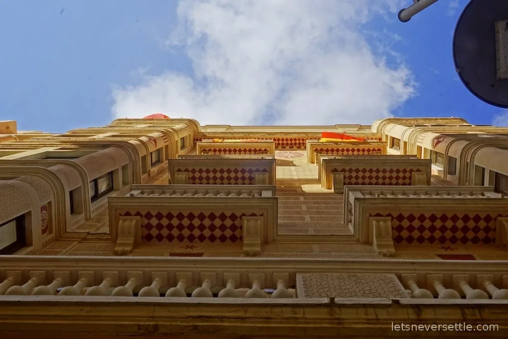 Looking straight up an ornate facade with red diamond-patterned balconies and Spanish flags