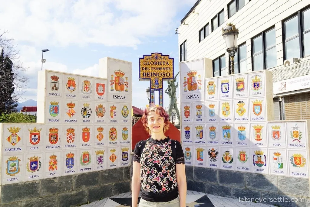 Inga standing before the tiled walls of the Glorieta del Teniente Reinoso, covered in the coats of arms of Spanish provinces