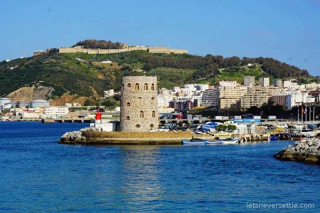 An old stone watchtower at the fishing harbour with small boats, and a hilltop fortress on the ridge behind