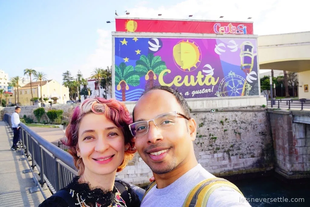 Inga and Dileep taking a selfie on the bridge over the Royal Walls moat, with the colourful Ceuta mural behind