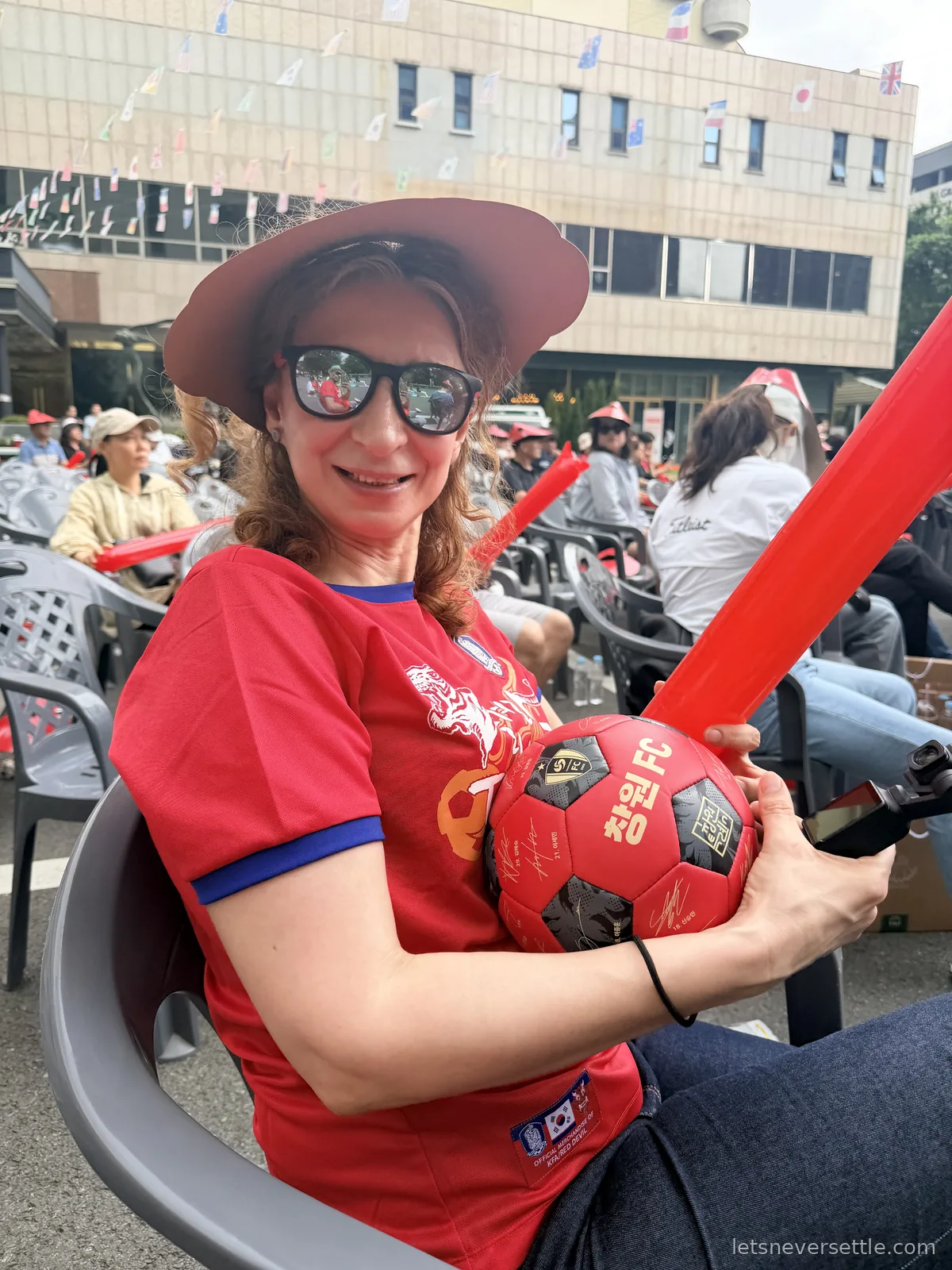 Inga in a red Korea shirt and wide red visor sitting at an outdoor watch party holding a signed Changwon FC football and a red inflatable clapper