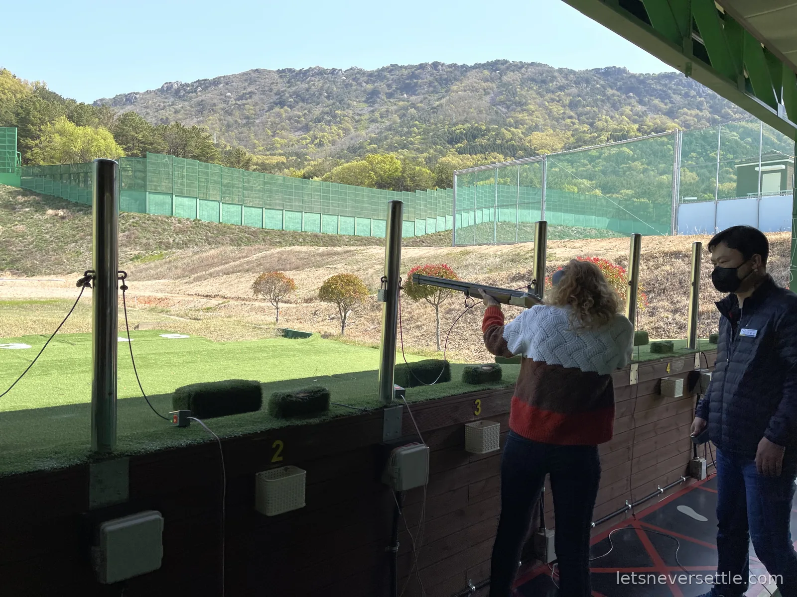Inga aiming a shotgun down a clay shooting range in Changwon while a masked instructor watches, wooded hills beyond