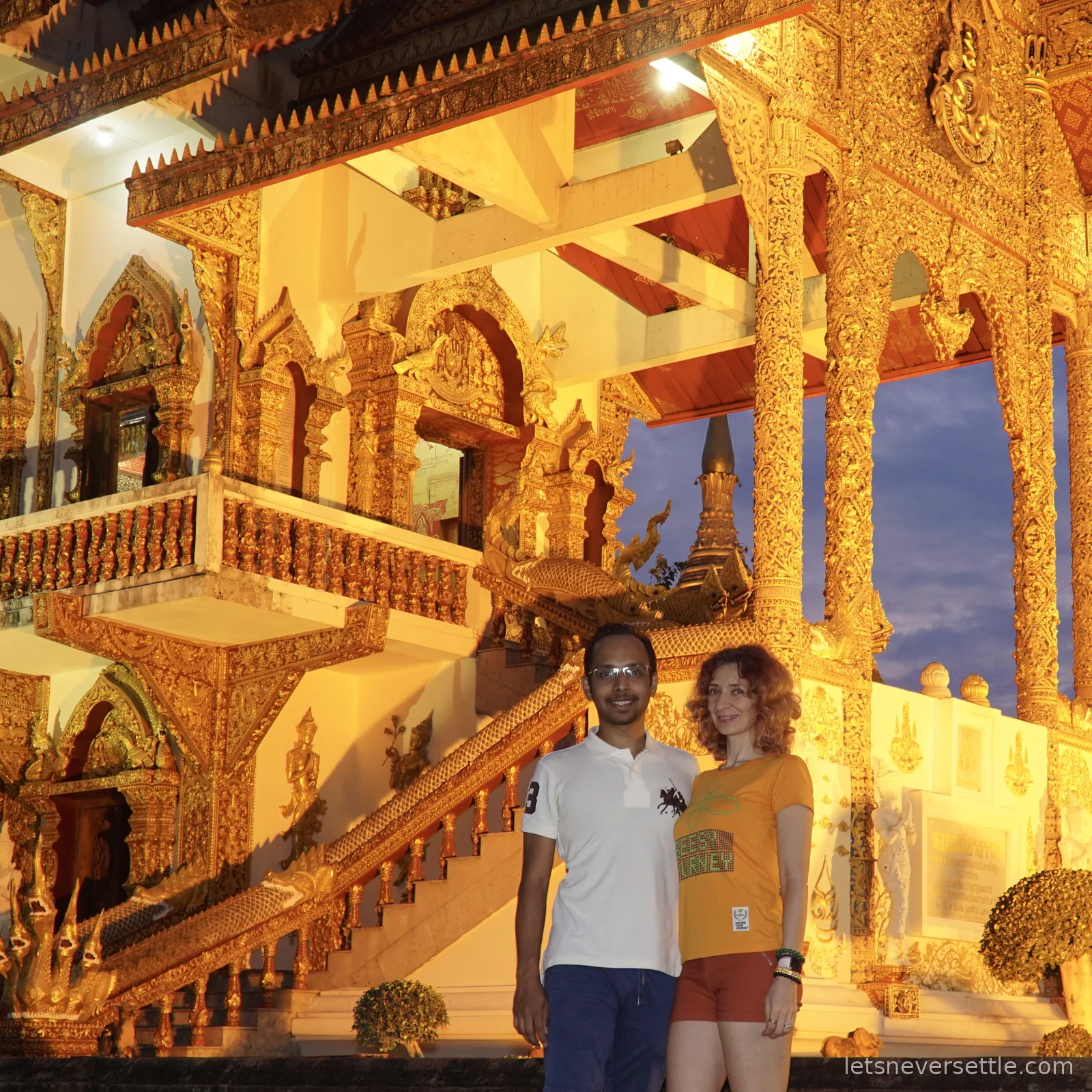 Inga and Dileep standing in front of a heavily gilded temple building with carved gold pillars, a staircase and a golden chedi spire rising behind it against a deep blue dusk sky