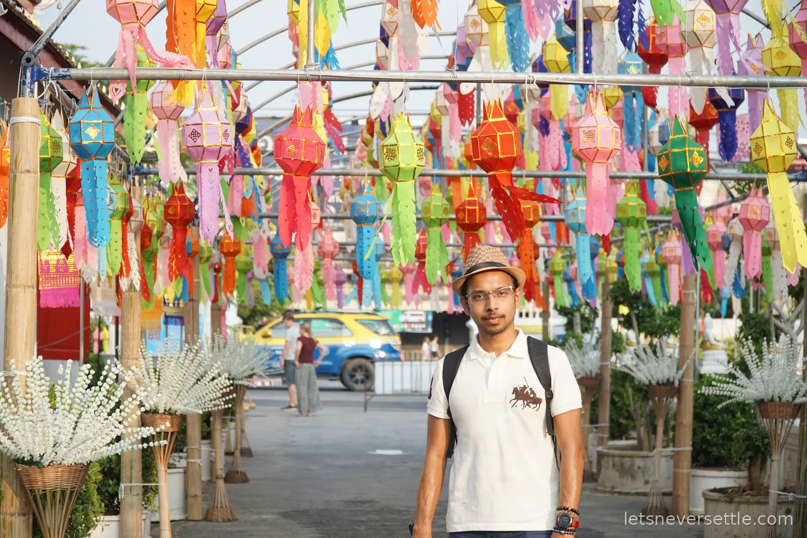 Dileep in a hat and white polo shirt standing under an arched walkway hung with hundreds of small multicoloured paper lanterns and cut-paper streamers