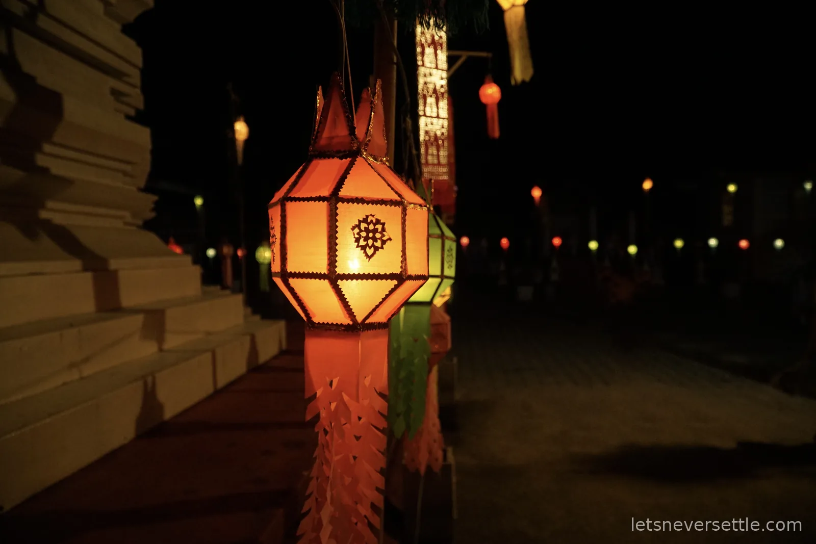 A glowing orange paper lantern hanging beside the stepped brick and plaster base of a temple chedi at night, with a line of small coloured lights along the path beyond