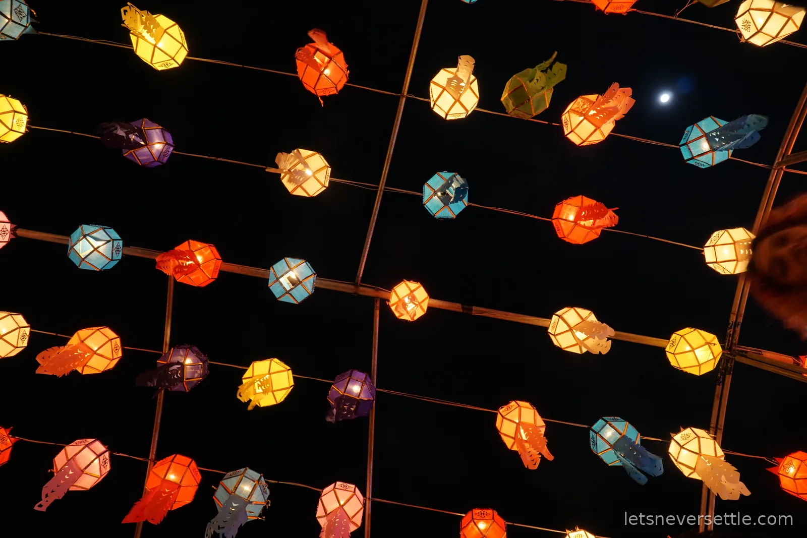 A grid of coloured paper lanterns strung on parallel wires against a black night sky, with the moon visible between them