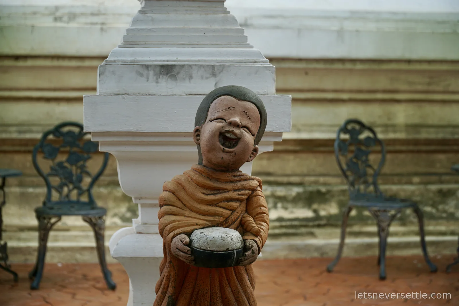 A terracotta statue of a laughing novice monk in orange robes holding an alms bowl, standing beside the white plaster base of a chedi with iron garden chairs behind