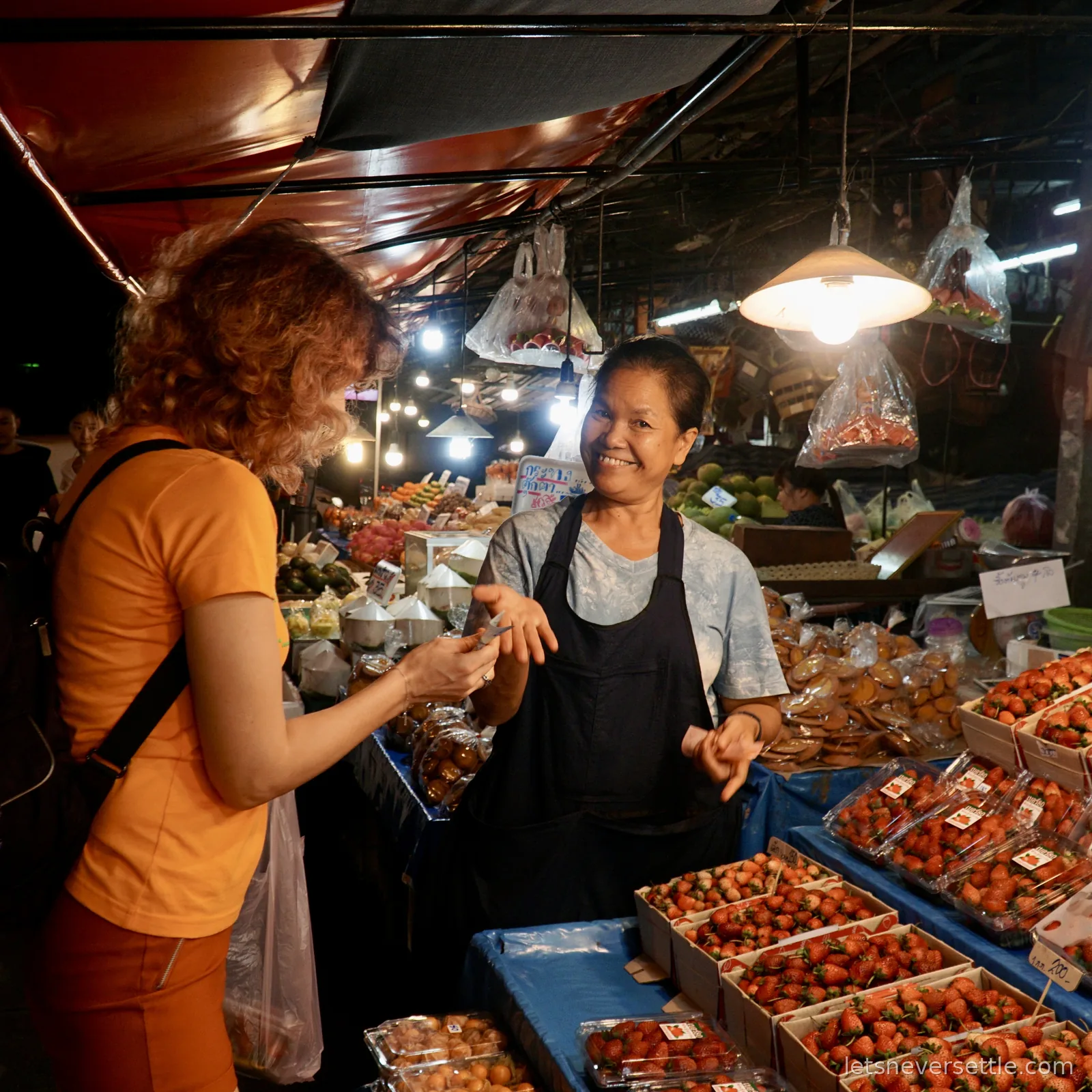 Inga talking to a smiling market vendor in an apron across a stall stacked with punnets of strawberries, tamarind and papaya under hanging bulbs at a Chiang Mai night market