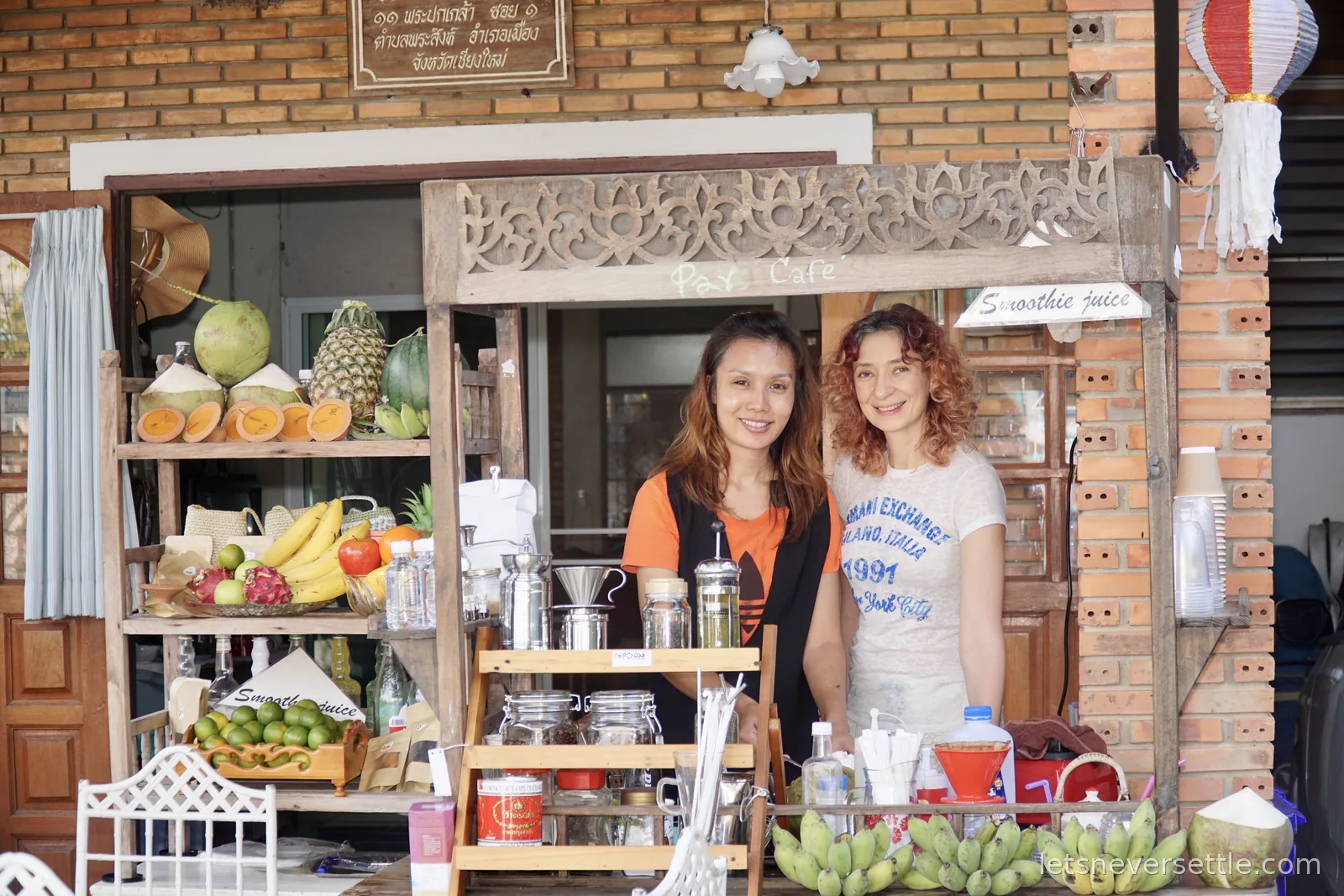 Inga and a Thai cafe owner standing behind a small carved wooden juice and coffee cart stacked with coconuts, pineapple, bananas and dragon fruit, under a Thai address sign for Phra Pokklao Soi 1