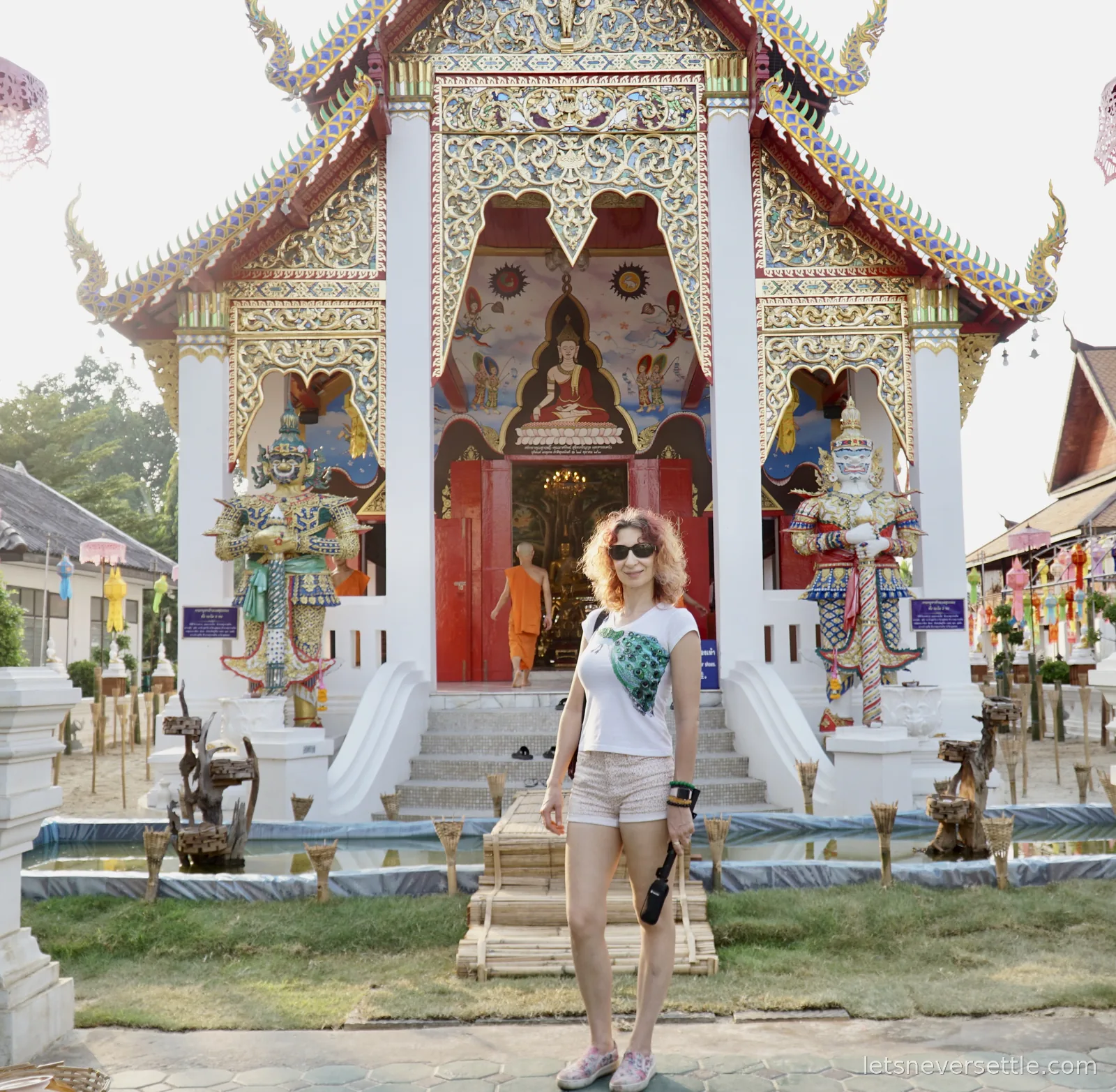 Inga standing in front of a white and gold temple viharn with a painted Buddha above the red doors, two ornate yaksha guardian statues flanking the steps and a monk in orange robes walking inside