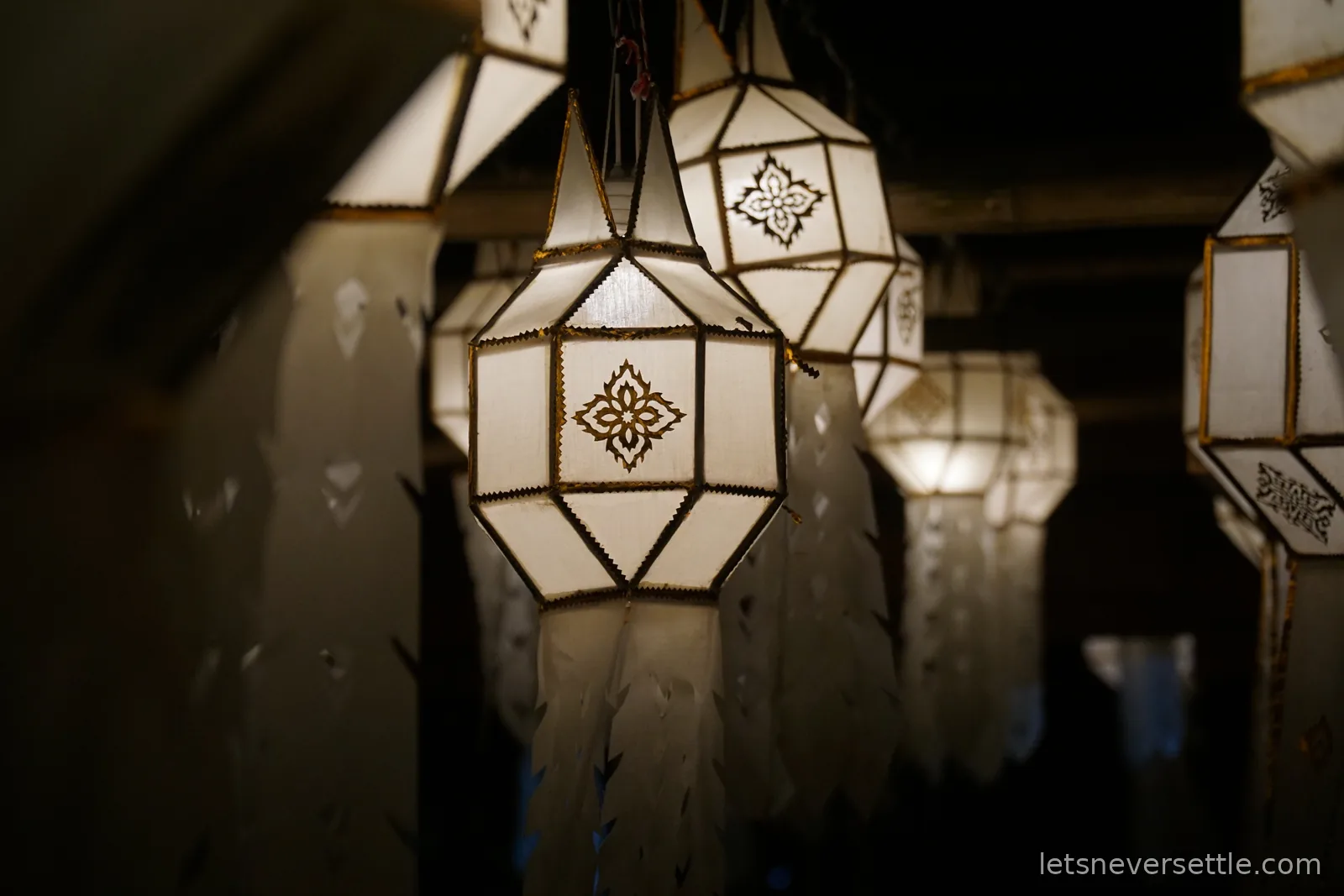 Close-up of white paper Lanna lanterns lit from within, each printed with a gold flower motif and hung with long cut-paper tails, receding into darkness under a wooden roof
