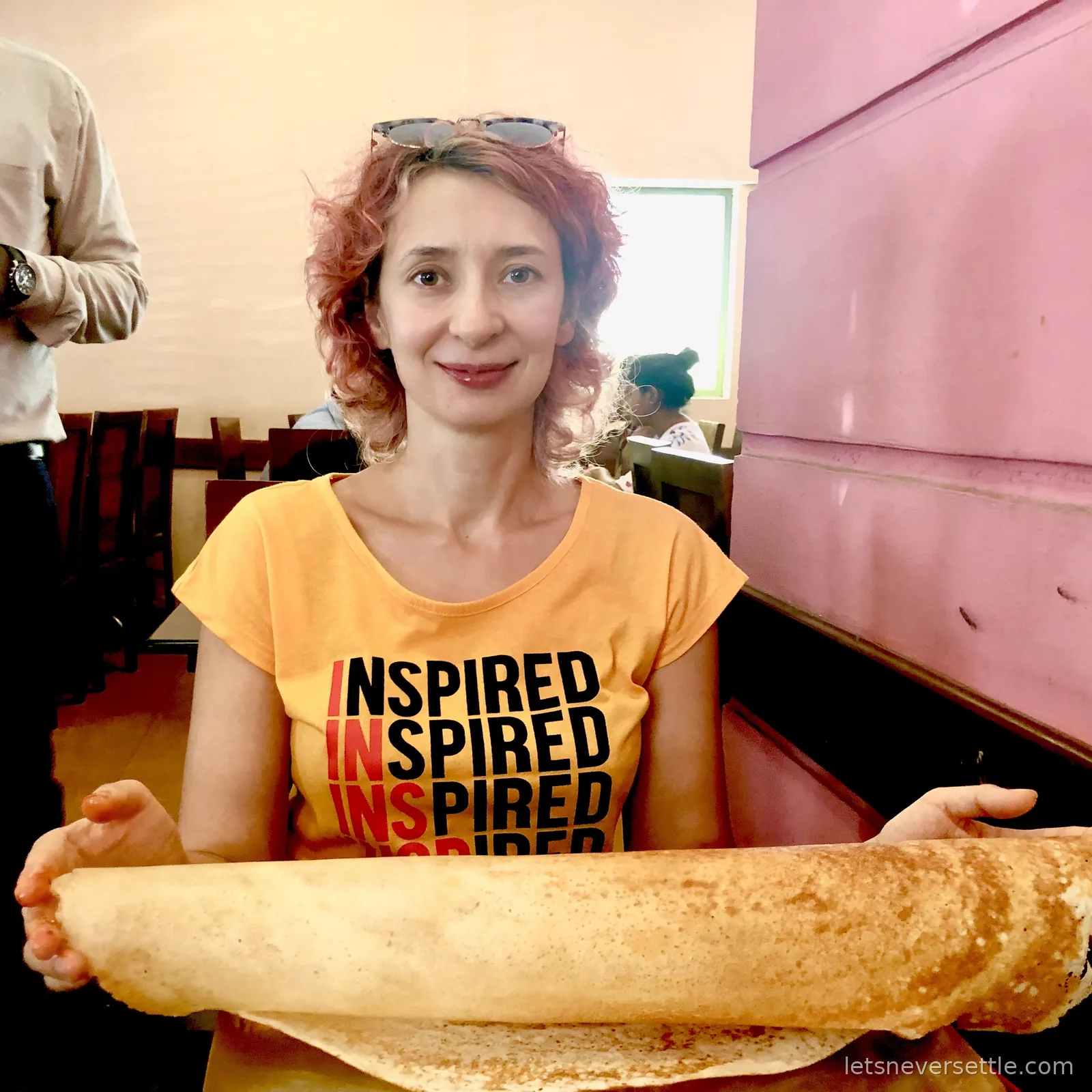 Inga in an orange t-shirt holding a huge rolled paper dosa across both hands at a plain cafe table, pink painted wall and wooden chairs behind her