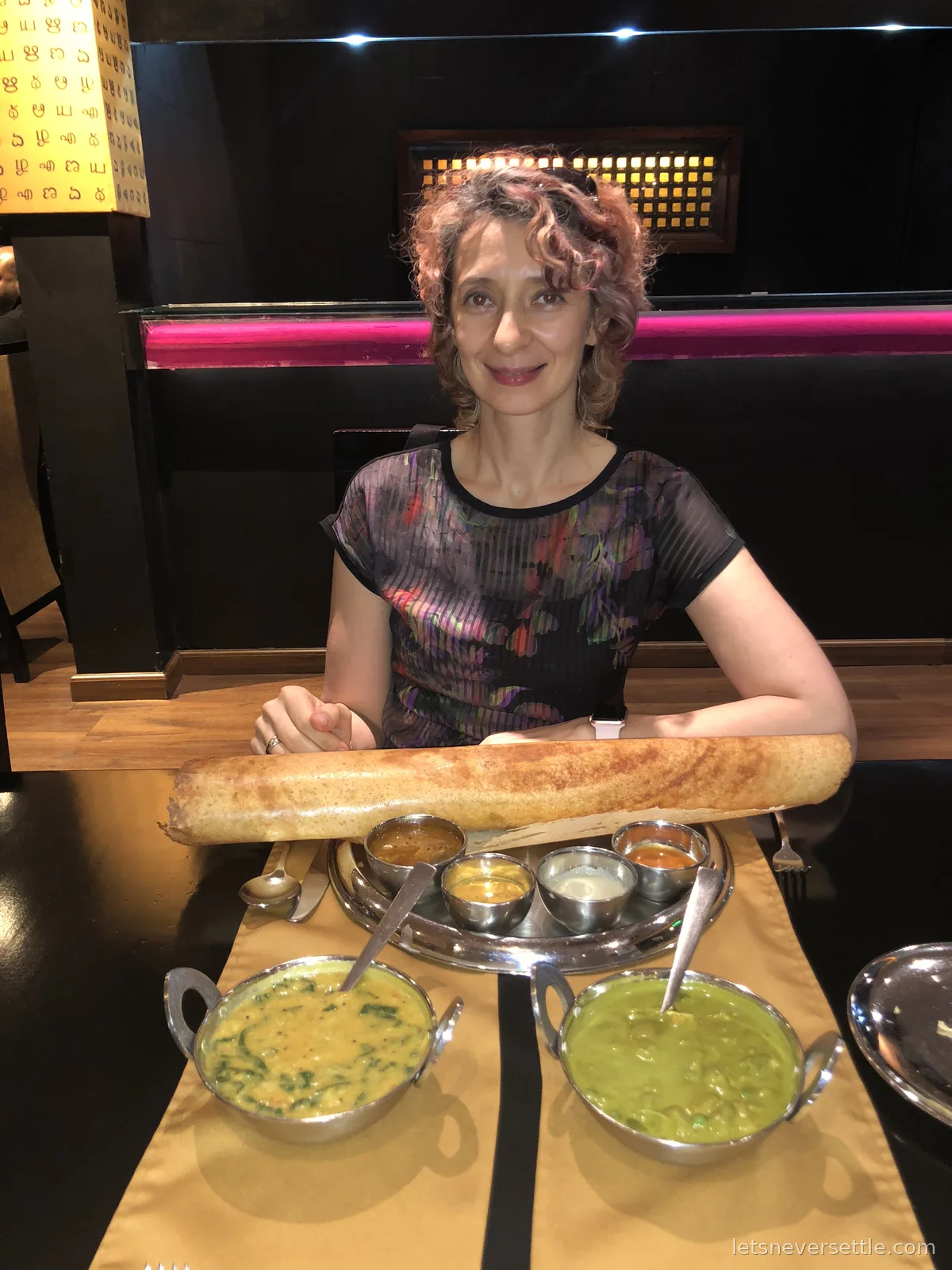 Inga at a restaurant table with a tall rolled paper dosa standing on a thali tray of steel cups with curries and chutneys