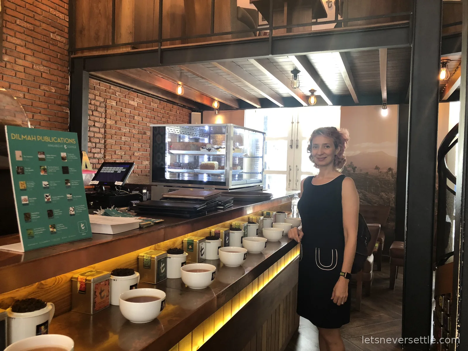 Inga standing at a Dilmah t-Lounge counter lined with white tasting bowls of brewed tea and colourful tea caddies