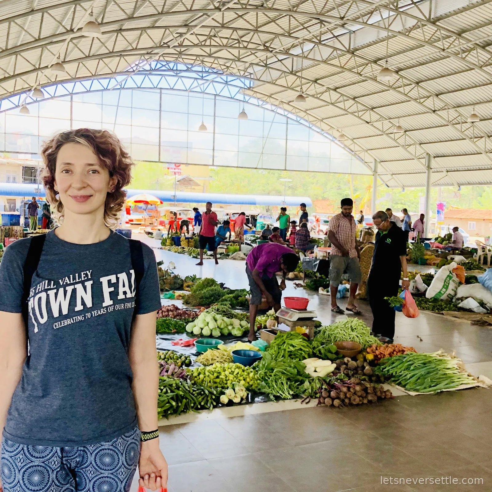 Inga standing in a covered vegetable market on the edge of Colombo, piles of okra, cabbages, greens and bananas laid out behind her