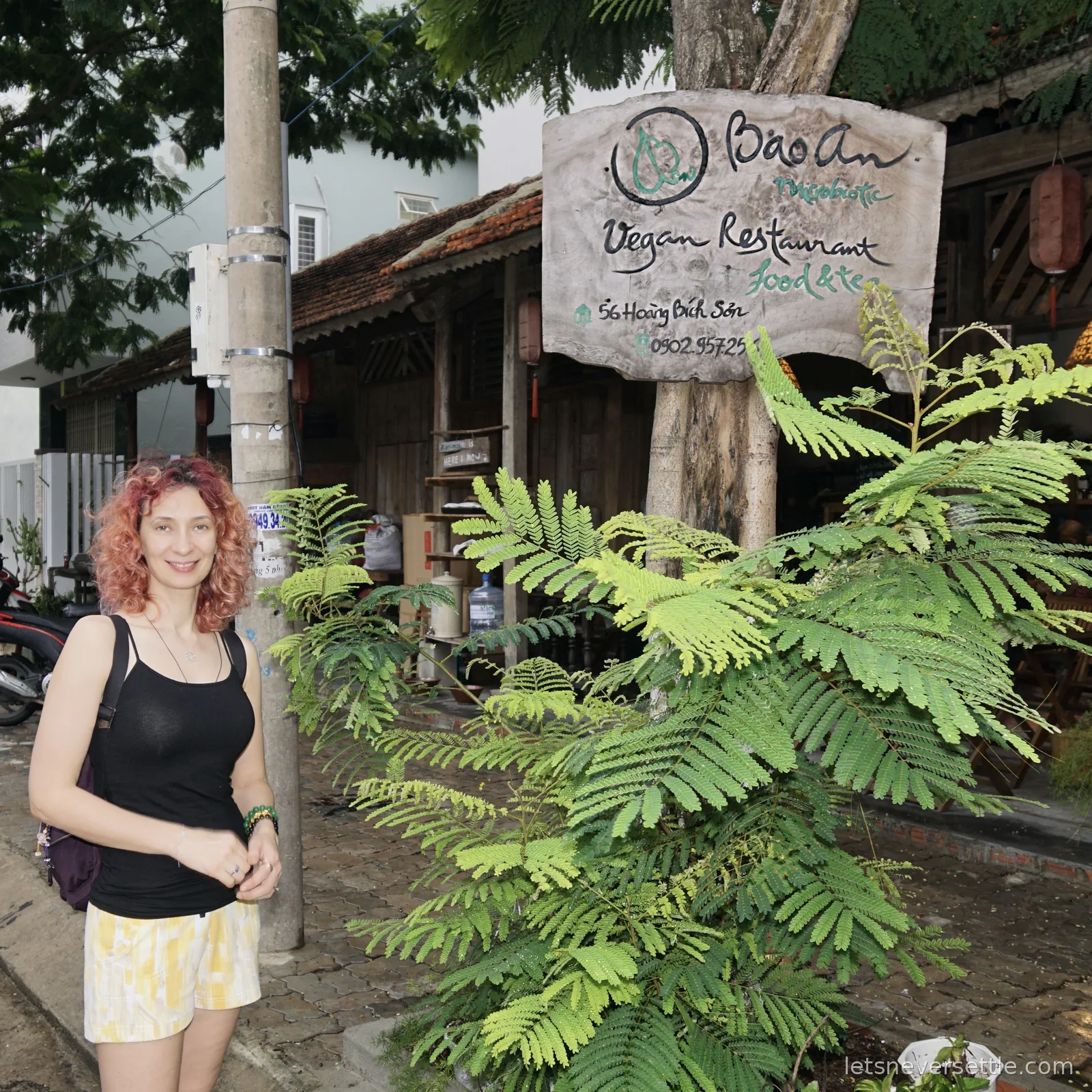 Inga standing on the pavement beside a hand-painted wooden sign reading Bao An Macrobiotic Vegan Restaurant, 56 Hoang Bich Son, in front of a low wooden-fronted building