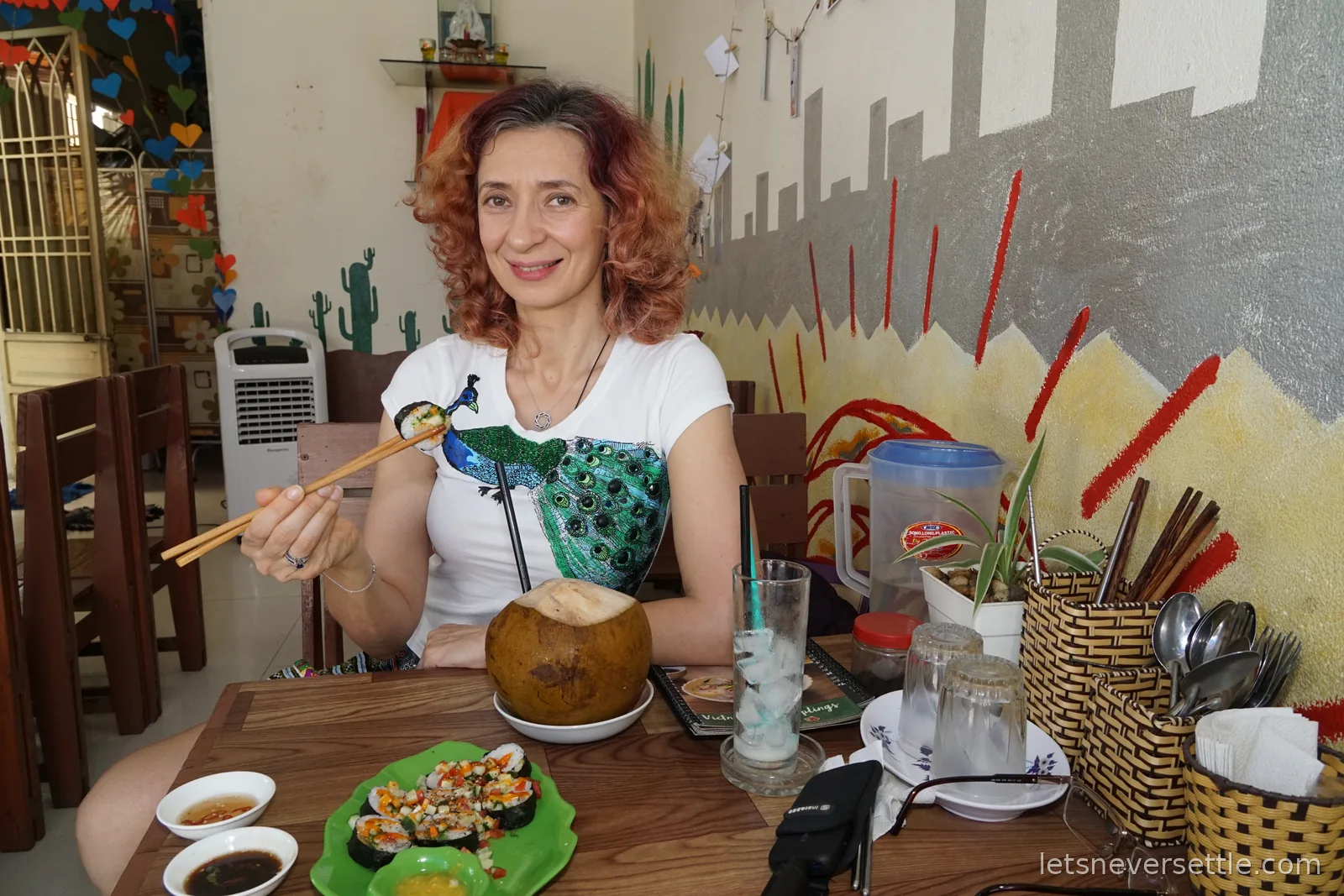 Inga holding chopsticks at a wooden table with a plate of Vietnamese sushi rolls and a whole young coconut, in front of a wall painted with cacti and a grey mountain mural