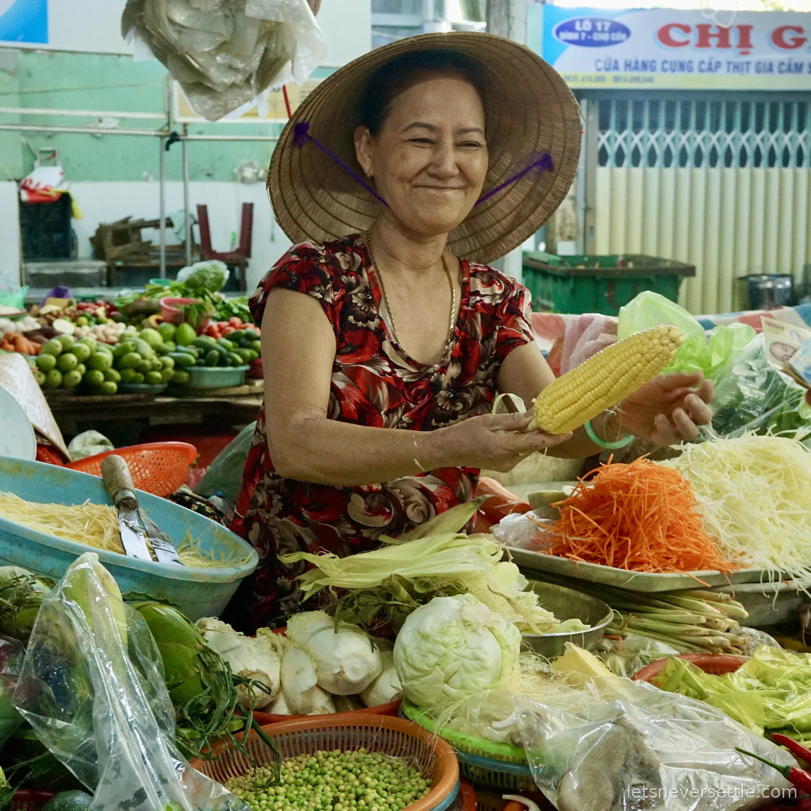 A smiling vendor in a conical hat holding up an ear of sweetcorn behind trays of shredded green papaya, carrot, beansprouts, cabbage and limes at Con Market in Da Nang
