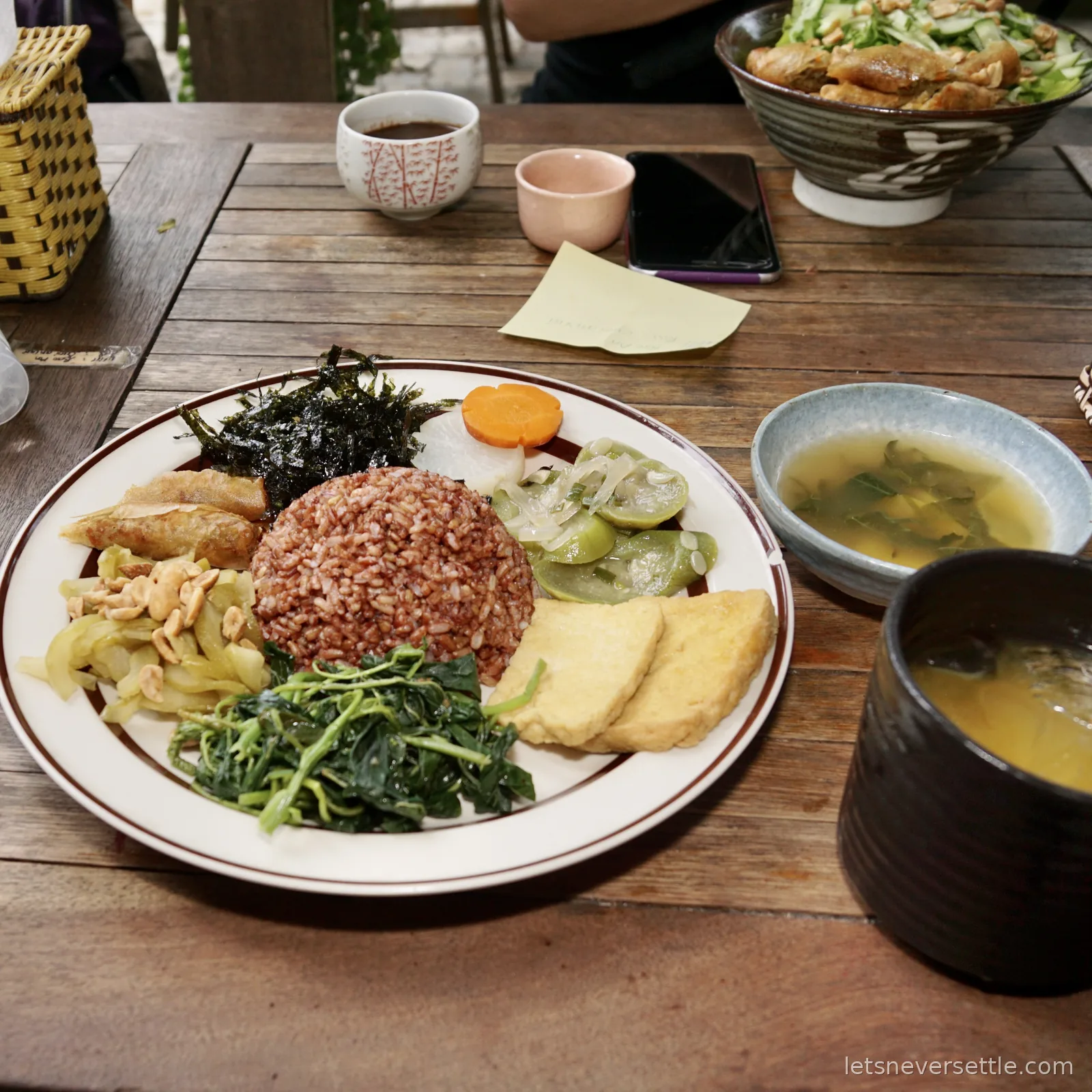 A macrobiotic set plate of brown rice, seaweed, wilted greens, fried tofu slices, pickled cucumber, cabbage with peanuts and carrot, beside a bowl of miso soup on a weathered wooden table