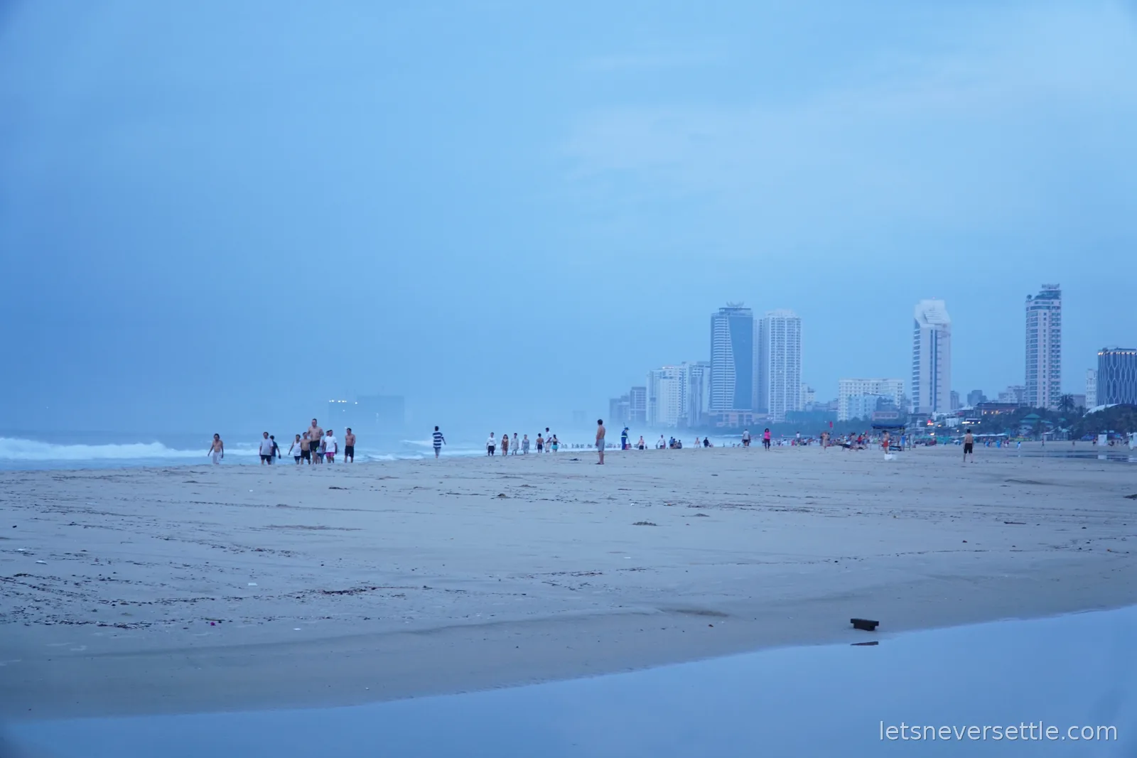 Wide empty sand at My Khe beach at dusk with swimmers in the surf and the pale high-rise towers of Da Nang along the shoreline behind