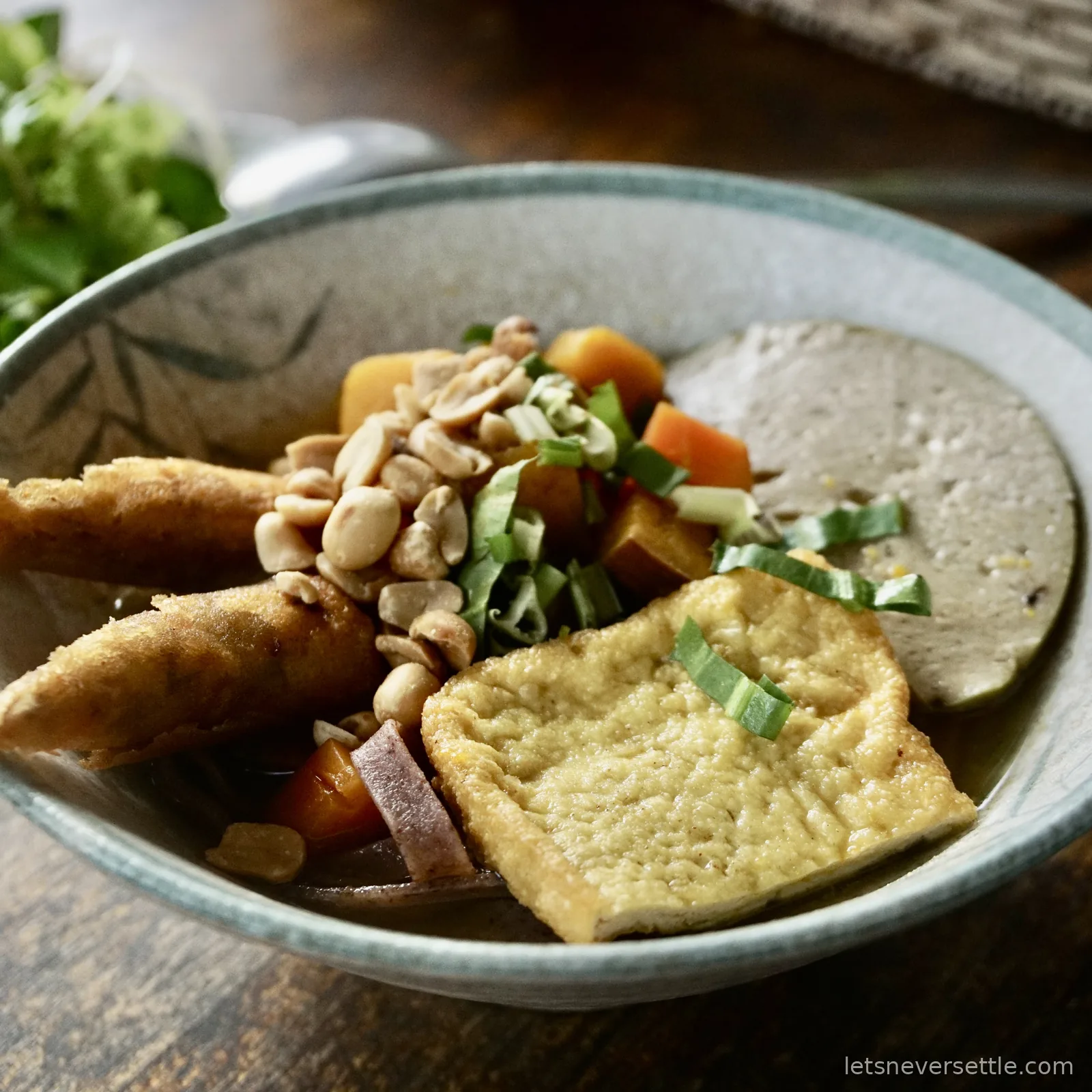 A close-up bowl of Vietnamese noodles topped with fried tofu, spring rolls, roasted peanuts, sliced mock ham, sweet potato, carrot and chopped spring onion