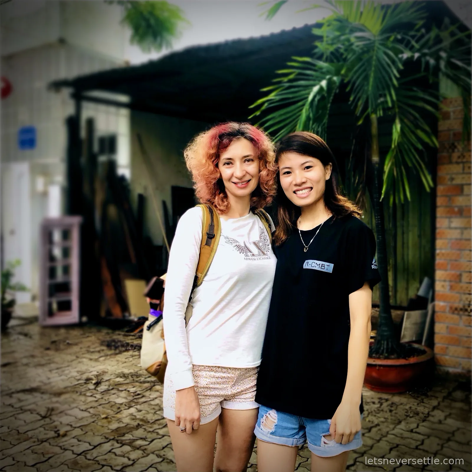 Inga standing arm in arm with a smiling young Vietnamese woman on a tiled courtyard, with a palm tree and a brick wall behind them