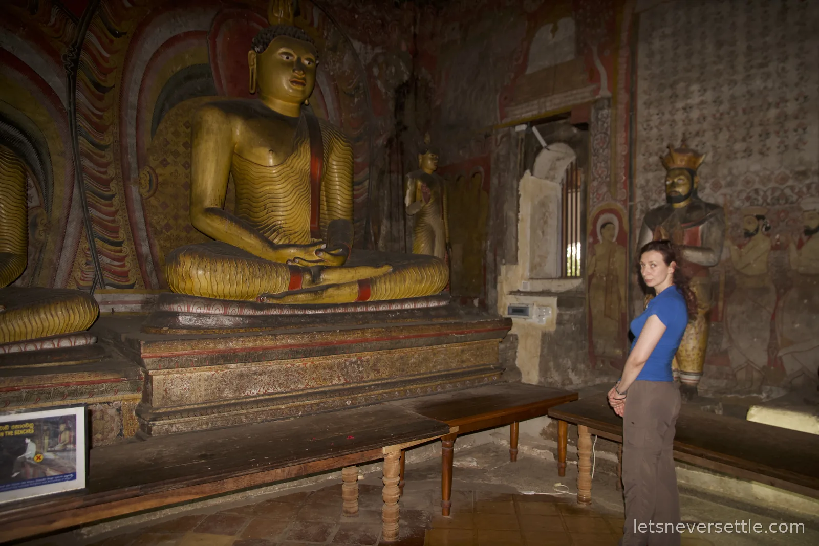 Inga inside a Dambulla cave beside a large golden seated Buddha, with a painted royal statue and murals covering the walls