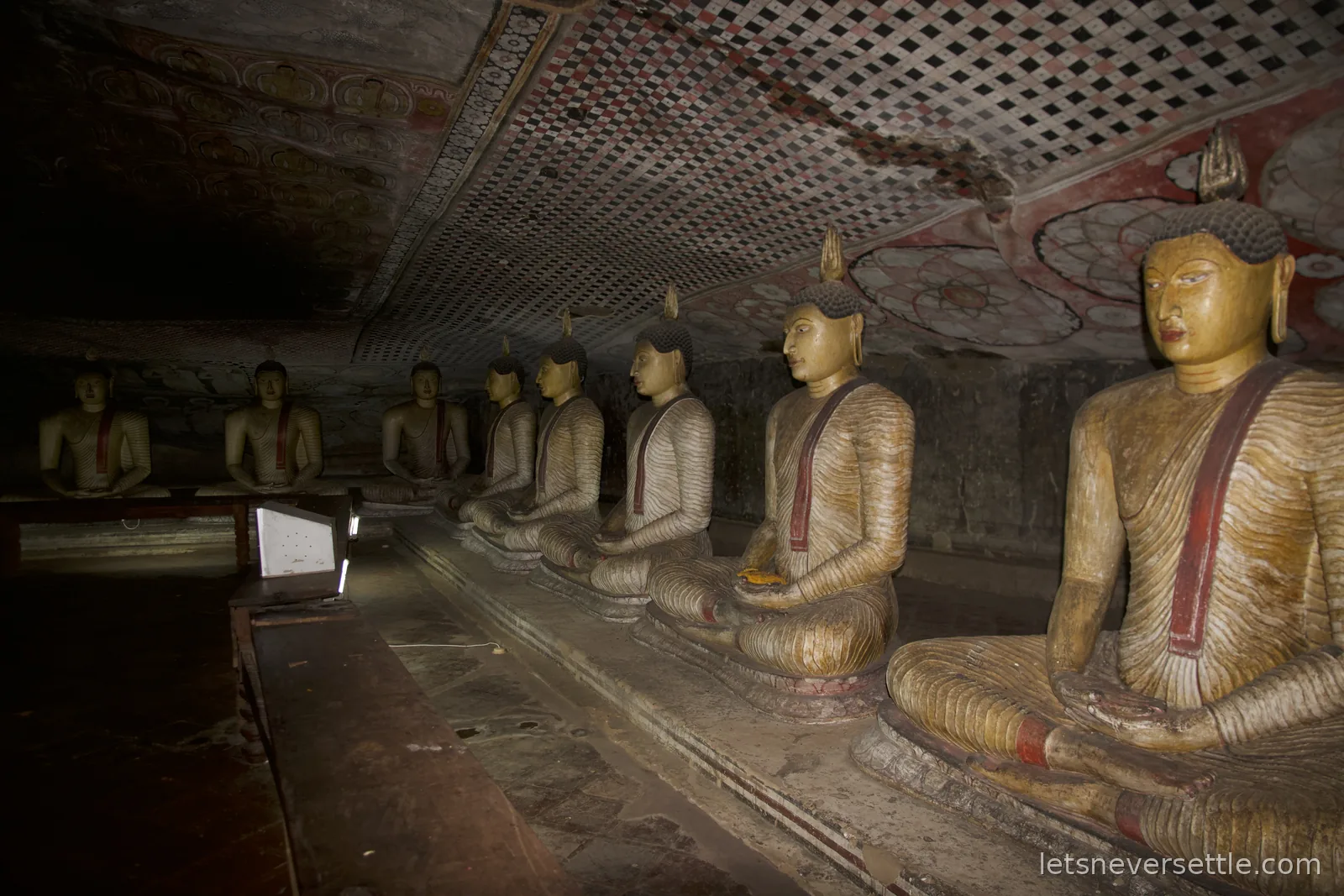 A long row of seated Buddha statues receding into a dim painted cave at Dambulla, the ceiling covered in chequered murals