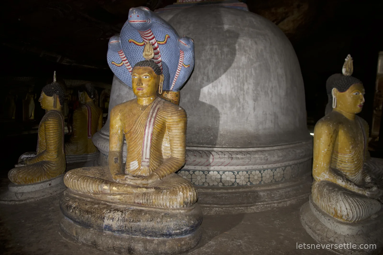 Seated Buddha statues around a stone stupa inside a Dambulla cave, one sheltered by a painted cobra hood