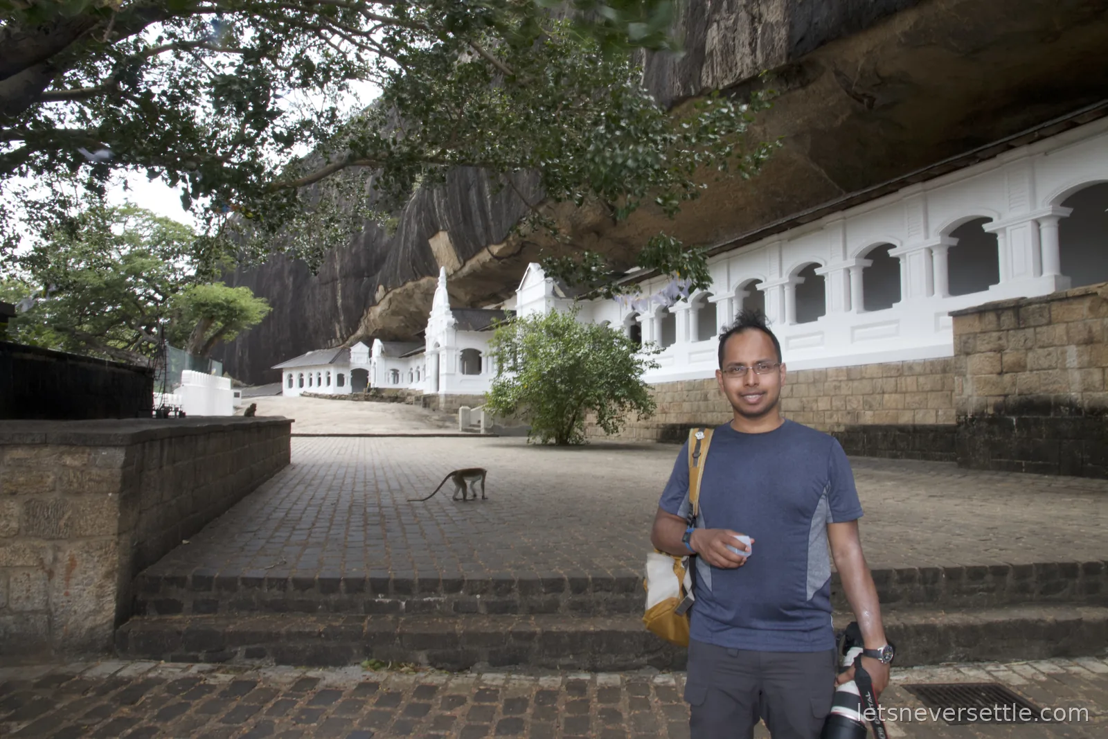 Dileep on the paved terrace outside the white colonnaded facade of the Dambulla cave temples, a monkey crossing the stones behind him