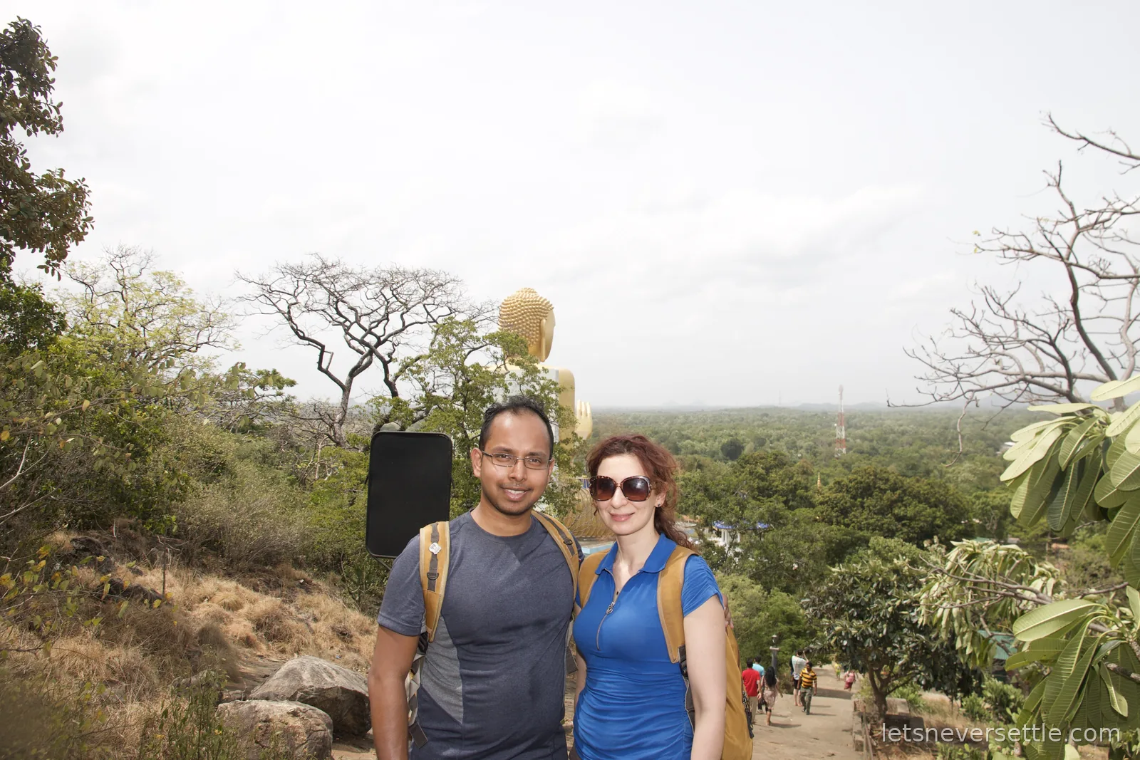 Dileep and Inga on the rock path at Dambulla with the giant golden seated Buddha of the Golden Temple behind them and green plains to the horizon